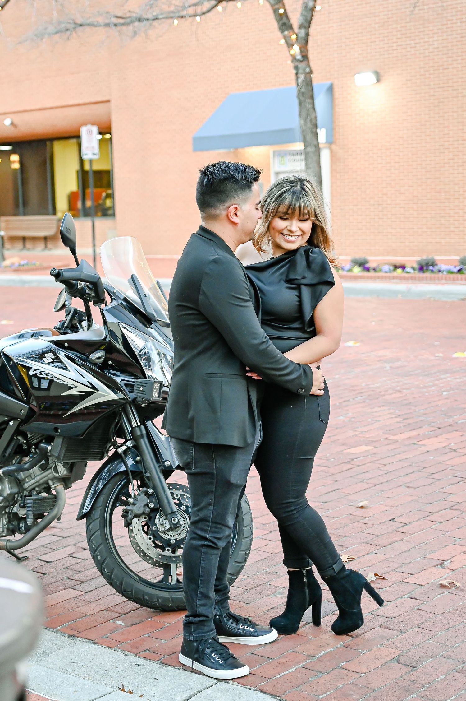 Couple in matching black outfits embracing near a motorcycle on a brick sidewalk.