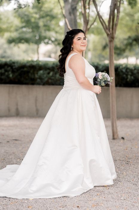 Smiling bride in white A-line wedding gown holds bouquet while standing among trees on gravel path.