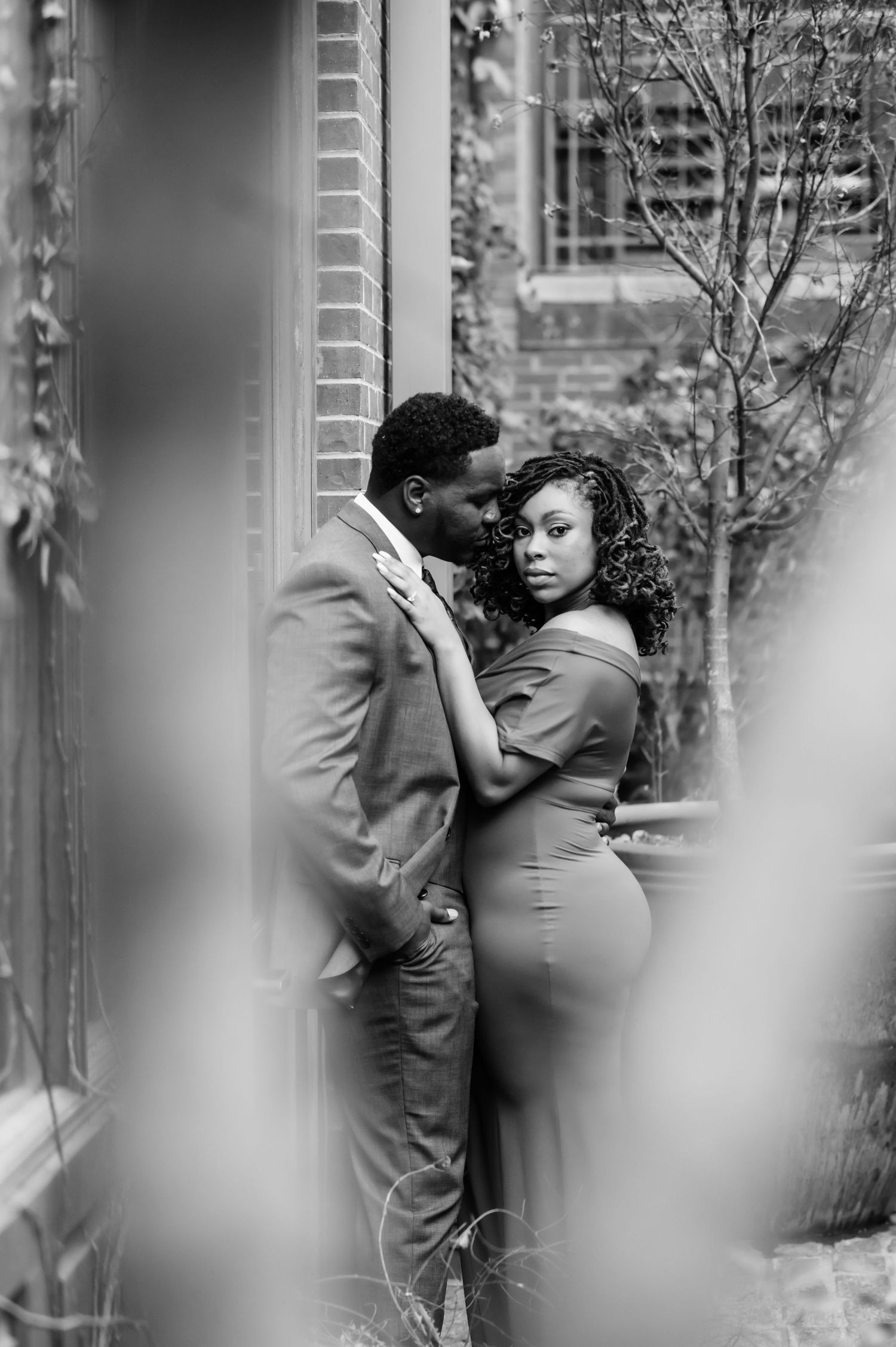 Black and white photo of a couple in formal wear sharing an intimate moment against a brick wall.
