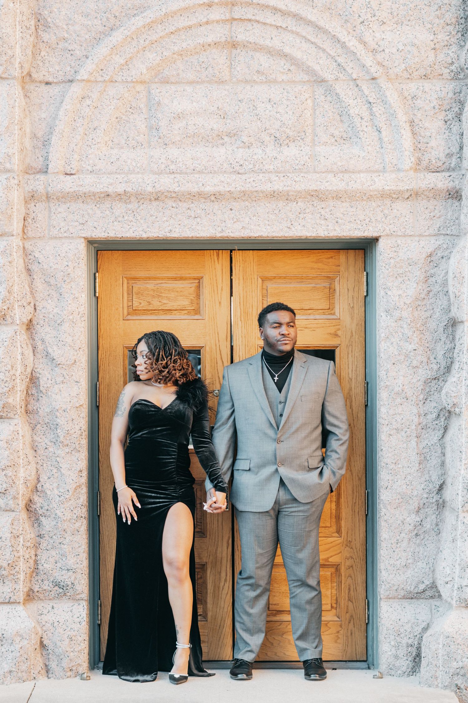 Couple in elegant attire posing before ornate wooden doors, woman in black velvet gown, man in gray suit holding hands.