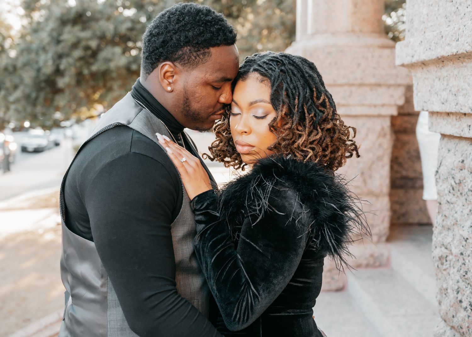 A couple shares an intimate embrace outdoors, dressed in elegant black attire near a stone column.