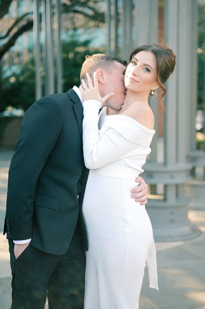 Groom in dark suit kisses bride's cheek as she smiles, wearing elegant off-shoulder white fitted gown.