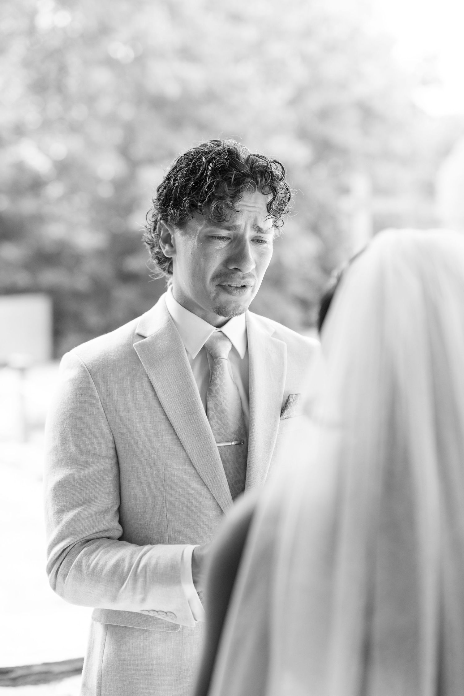 Groom in light suit looking emotional during outdoor wedding ceremony in black and white photo.