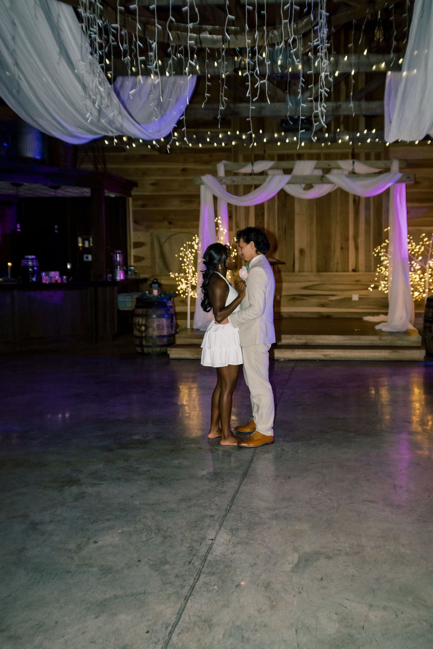 Couple sharing romantic first dance in rustic barn wedding venue decorated with string lights and white draping.