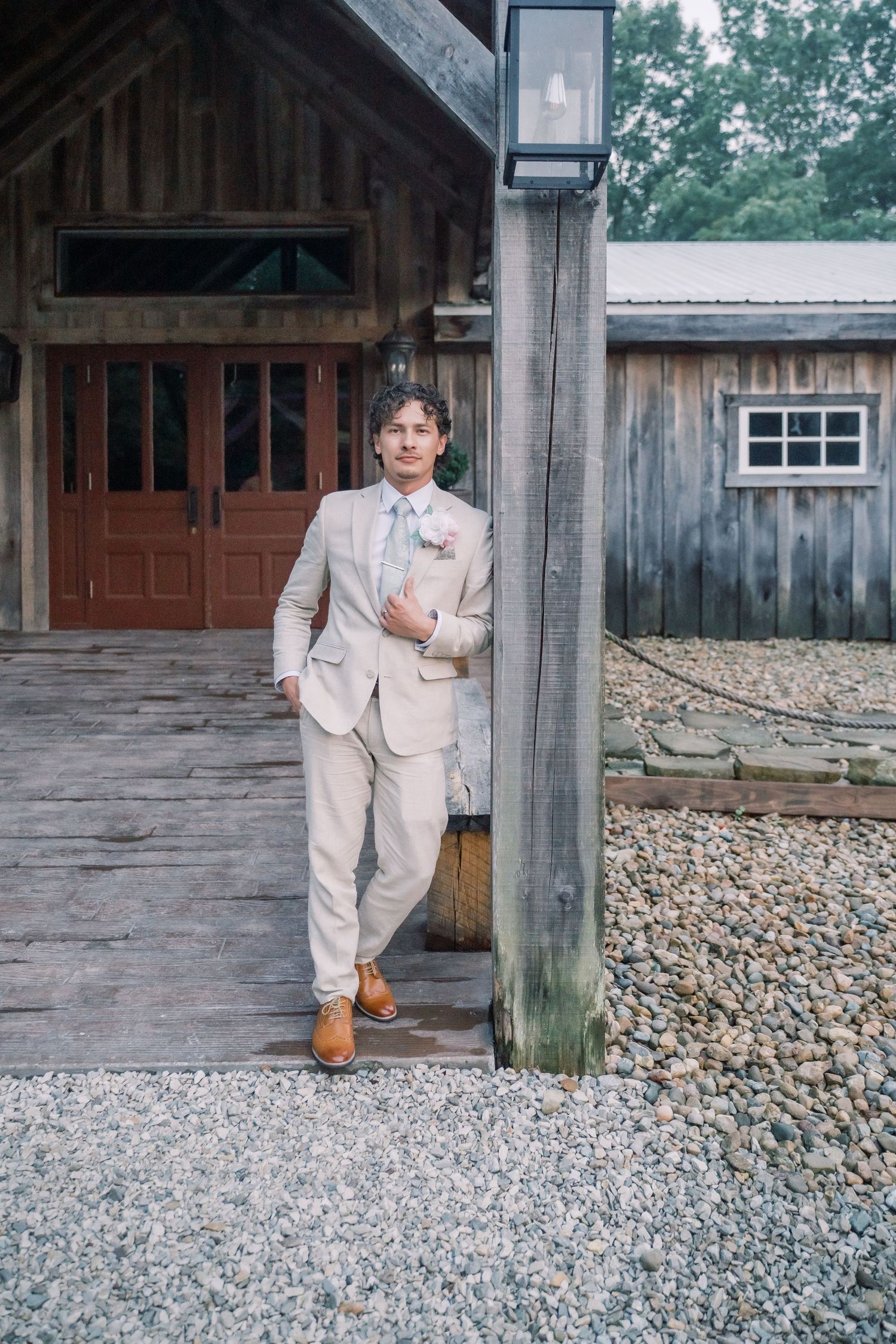 A groom in a beige suit and floral boutonniere leans against a wooden post outside a rustic barn venue.