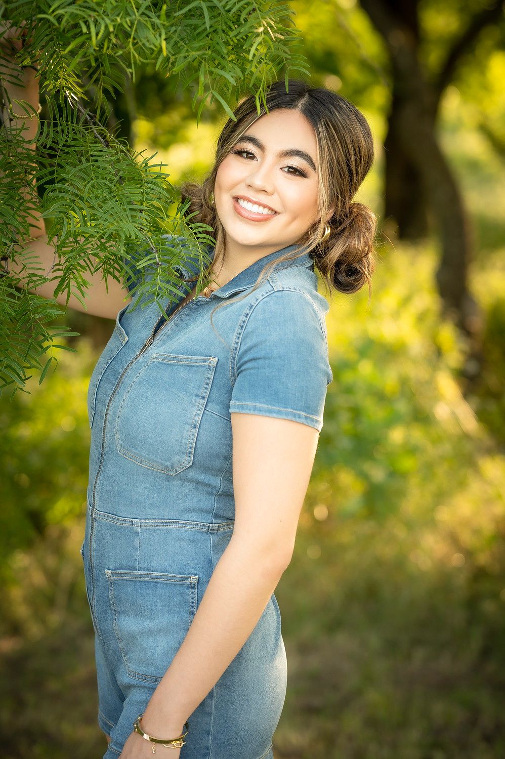 Young woman smiling in denim jumpsuit outdoors among green trees in warm sunlight.
