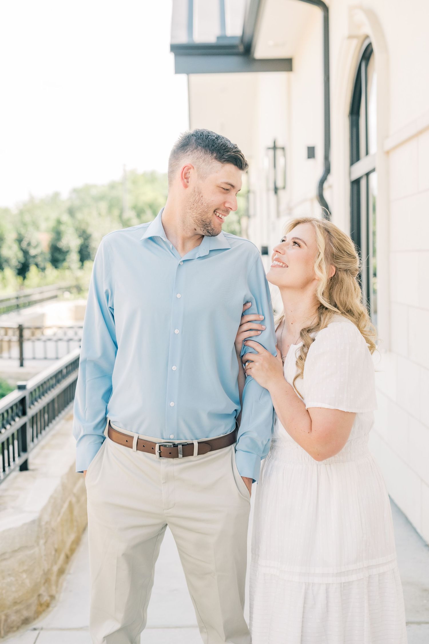 A couple in coordinating blue and white outfits smile at each other on a sunlit outdoor walkway.