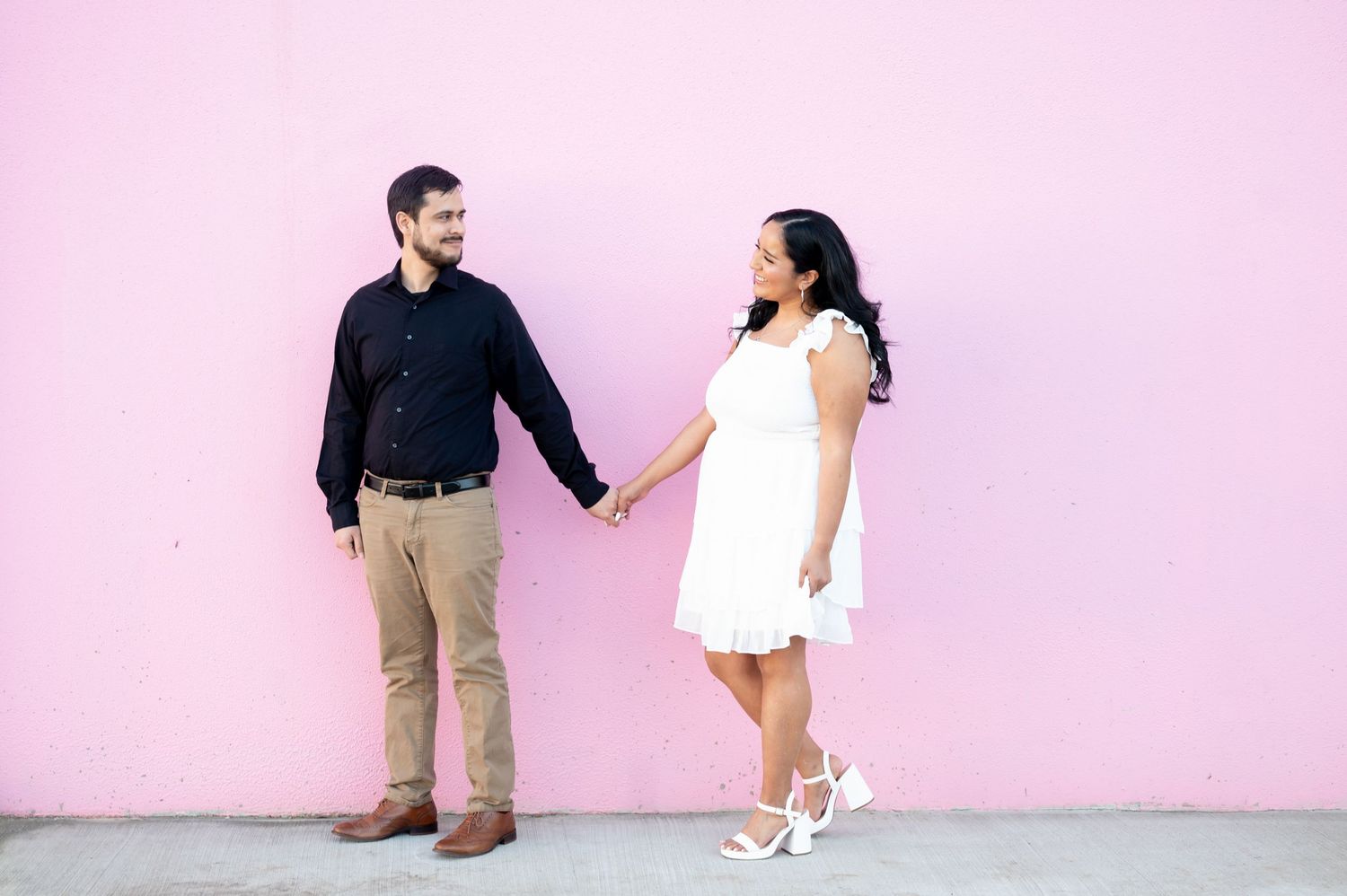 A couple holds hands while standing against a pink wall, with her wearing a white dress and him in dark clothing.