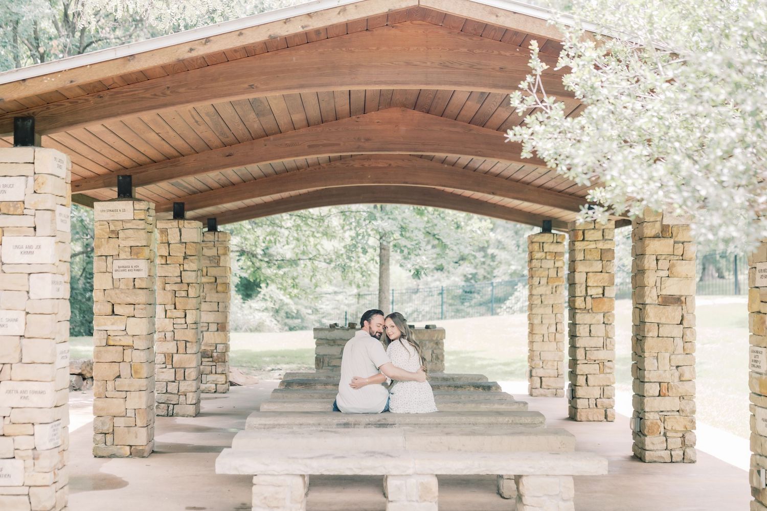 Couple sitting close together under a rustic stone and wood pavilion surrounded by lush greenery and blooming trees.
