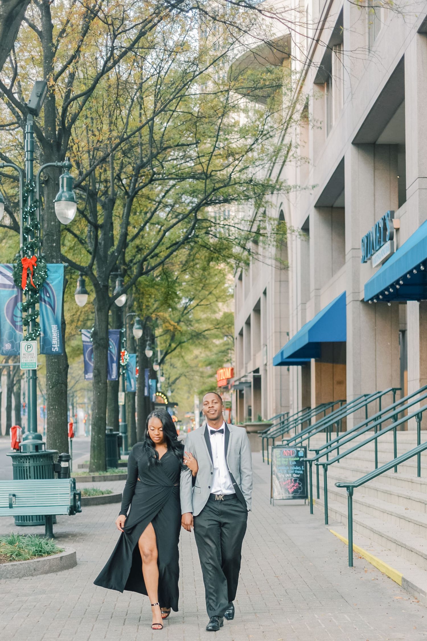 Couple holding hands in formal attire strolling along an urban sidewalk lined with autumn trees.