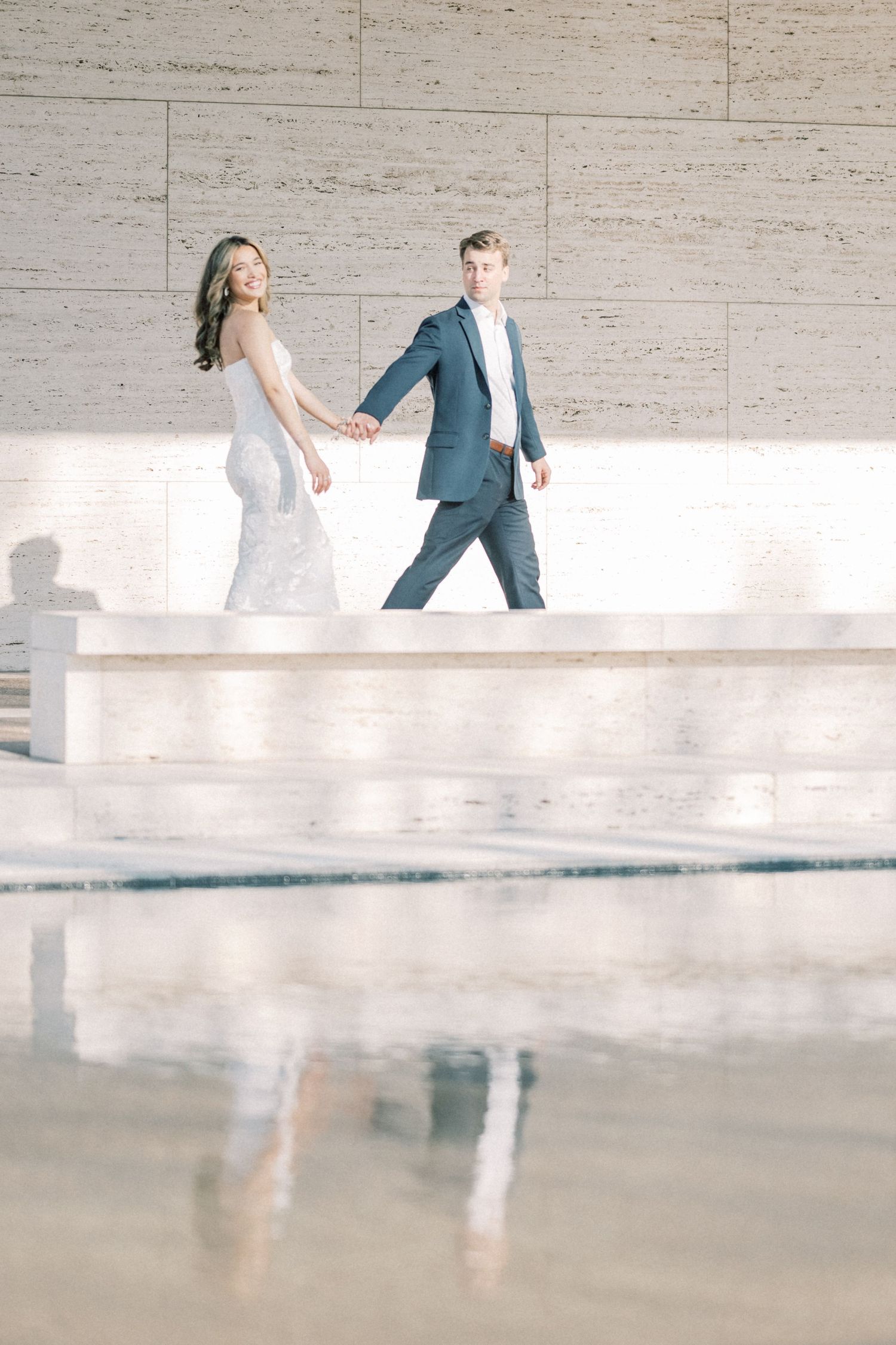 A couple in wedding attire walks together along a modern concrete wall with reflective flooring below.