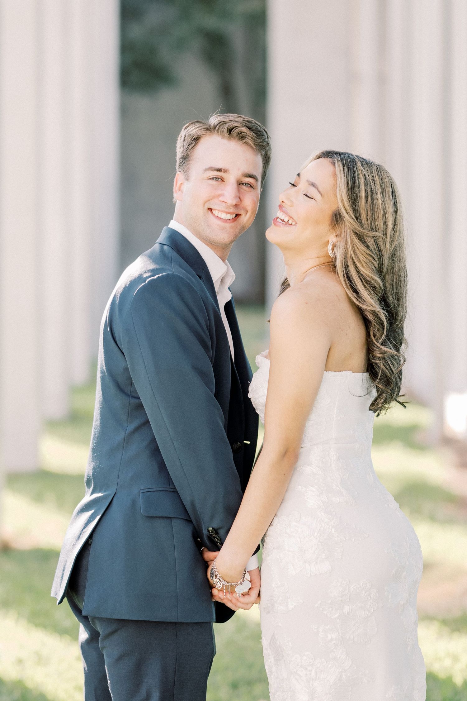 A joyful couple in wedding attire share a candid moment together while holding hands outdoors by elegant columns.