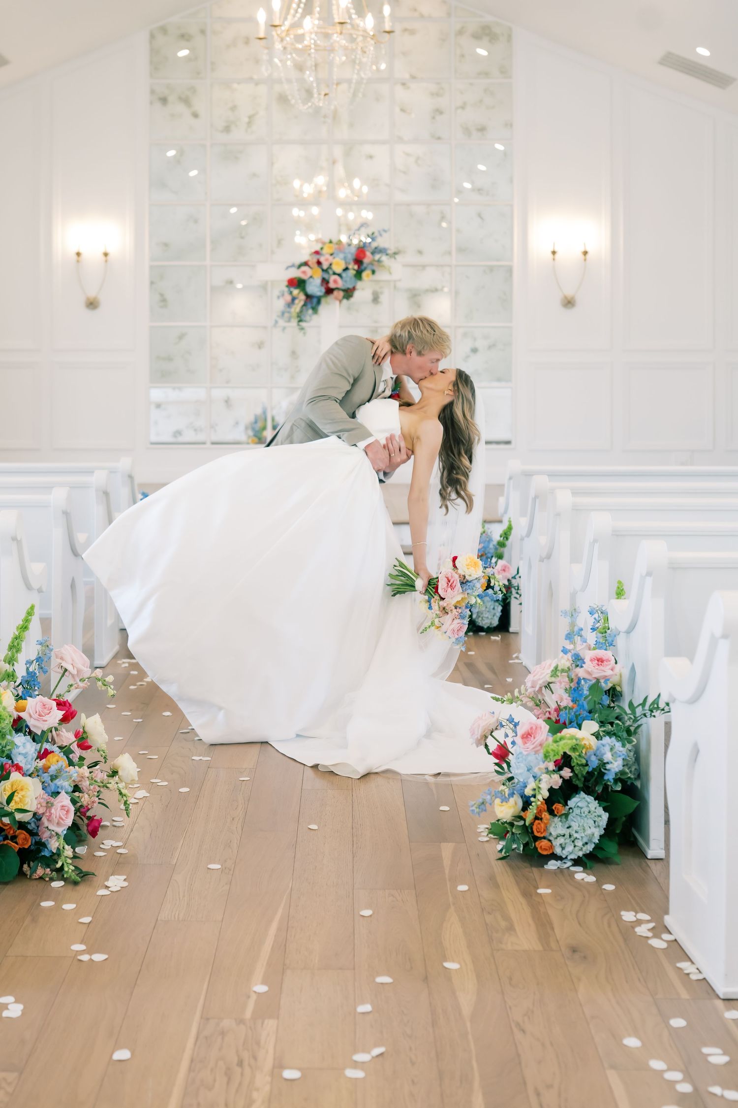 Groom dips bride for a kiss in a white chapel aisle lined with colorful floral arrangements.