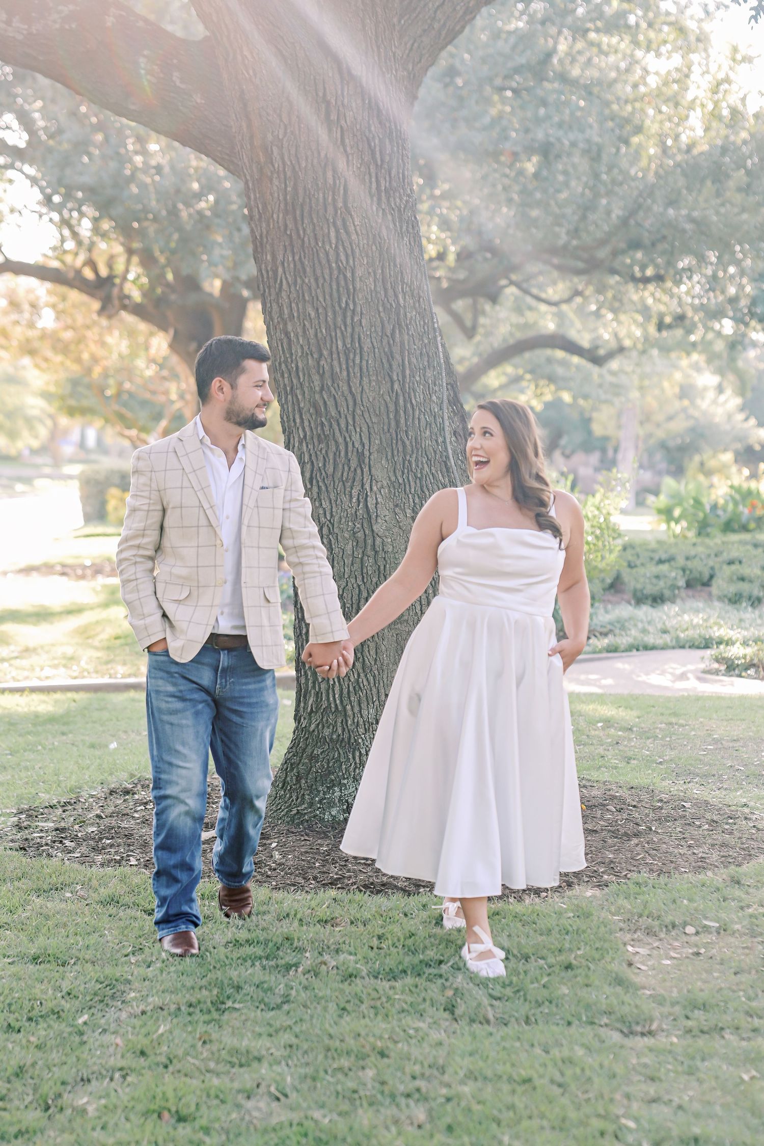 Engaged couple holds hands and walks toward the camera beneath a large oak tree, the woman laughing in a flowing white dress.