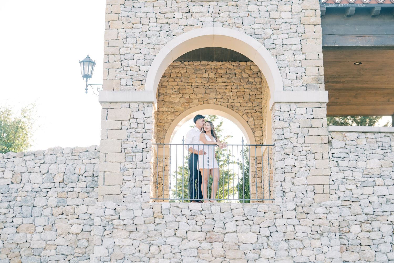 Couple sharing a romantic kiss under a stone arch on a rustic Mediterranean wall during an outdoor engagement session.
