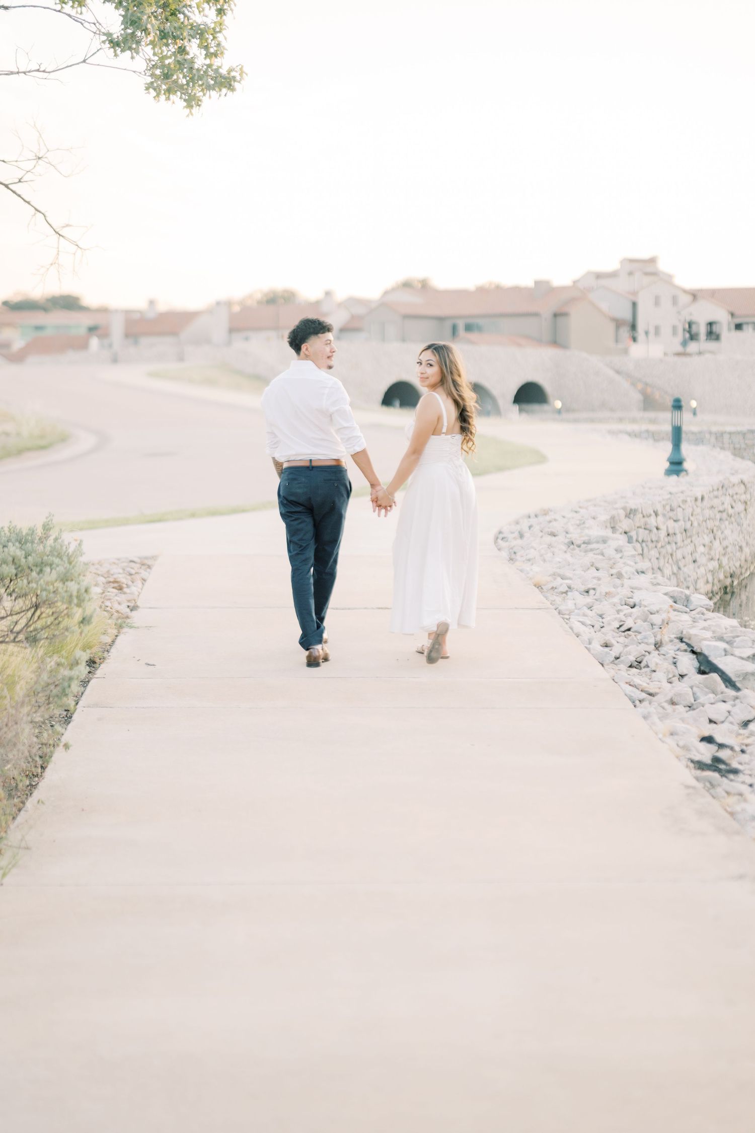 Couple walking hand in hand along scenic pathway during soft sunset engagement session.