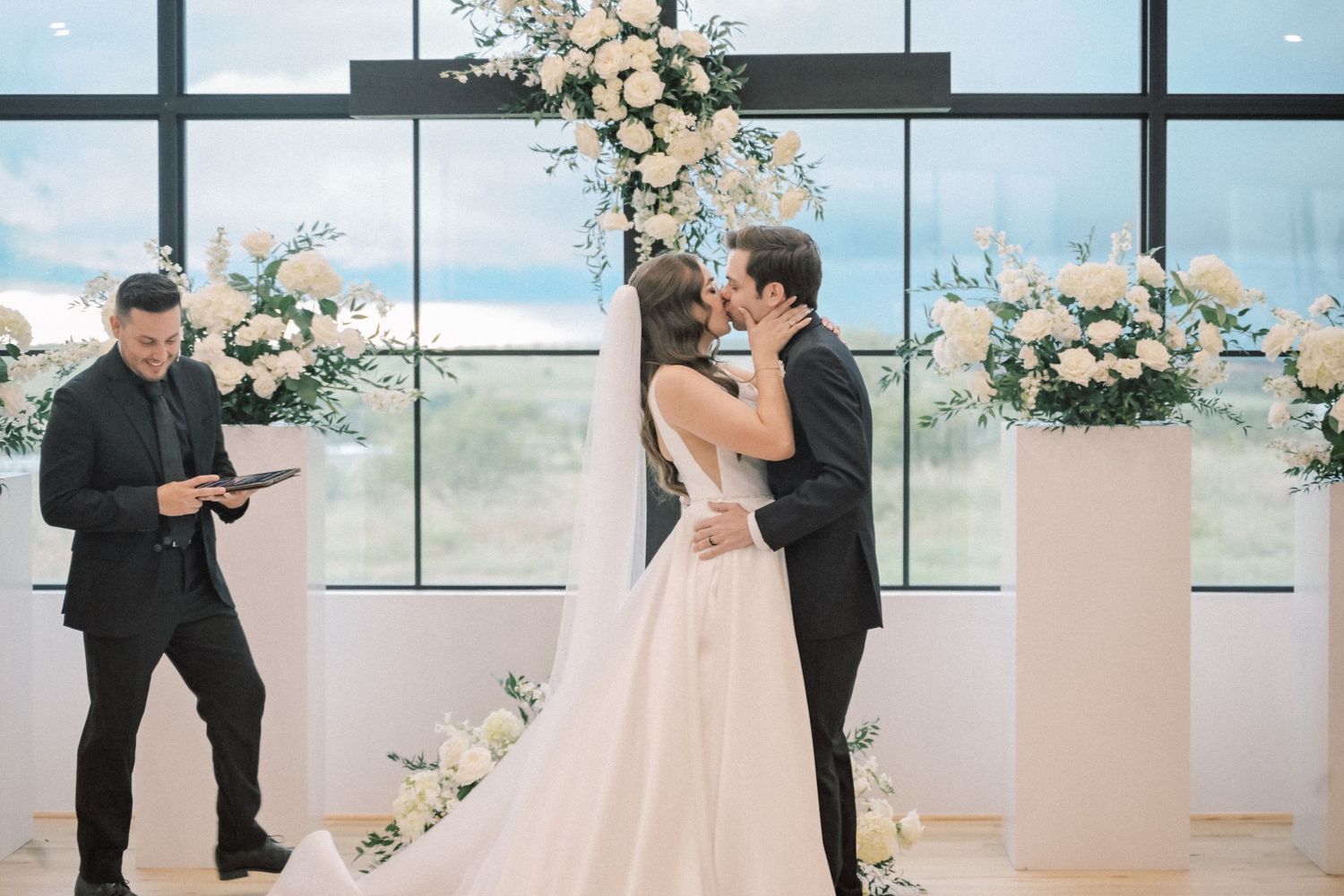 Bride and groom share first kiss at altar surrounded by white floral arrangements.