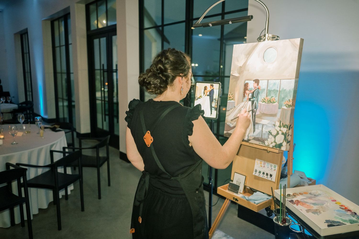 Artist painting live wedding portrait on canvas at easel during evening reception.