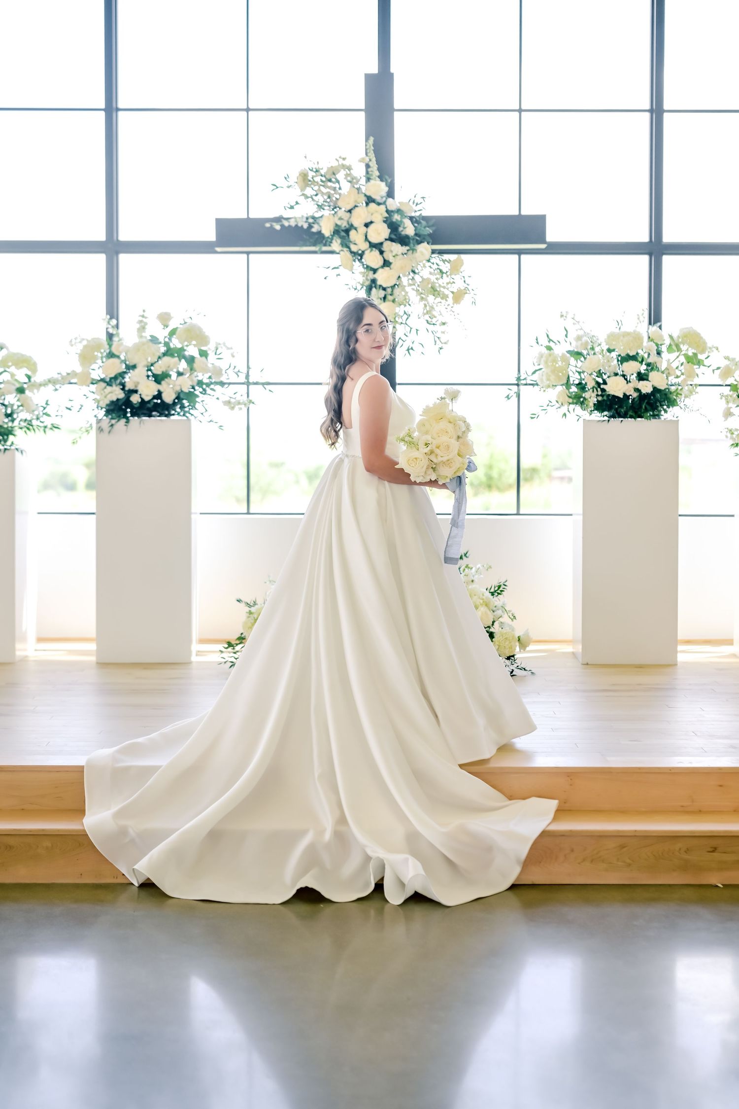 Bride in elegant white ball gown holding bouquet stands before floral arrangements in bright venue.