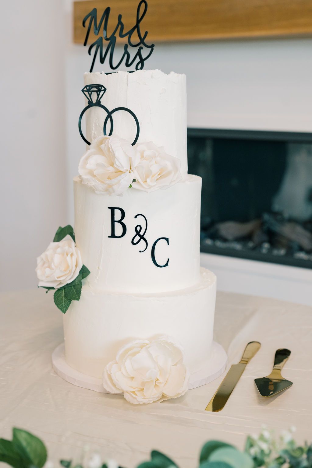 Elegant three-tier white wedding cake with B&C monogram, floral accents, ring design, and Mr & Mrs topper near fireplace.