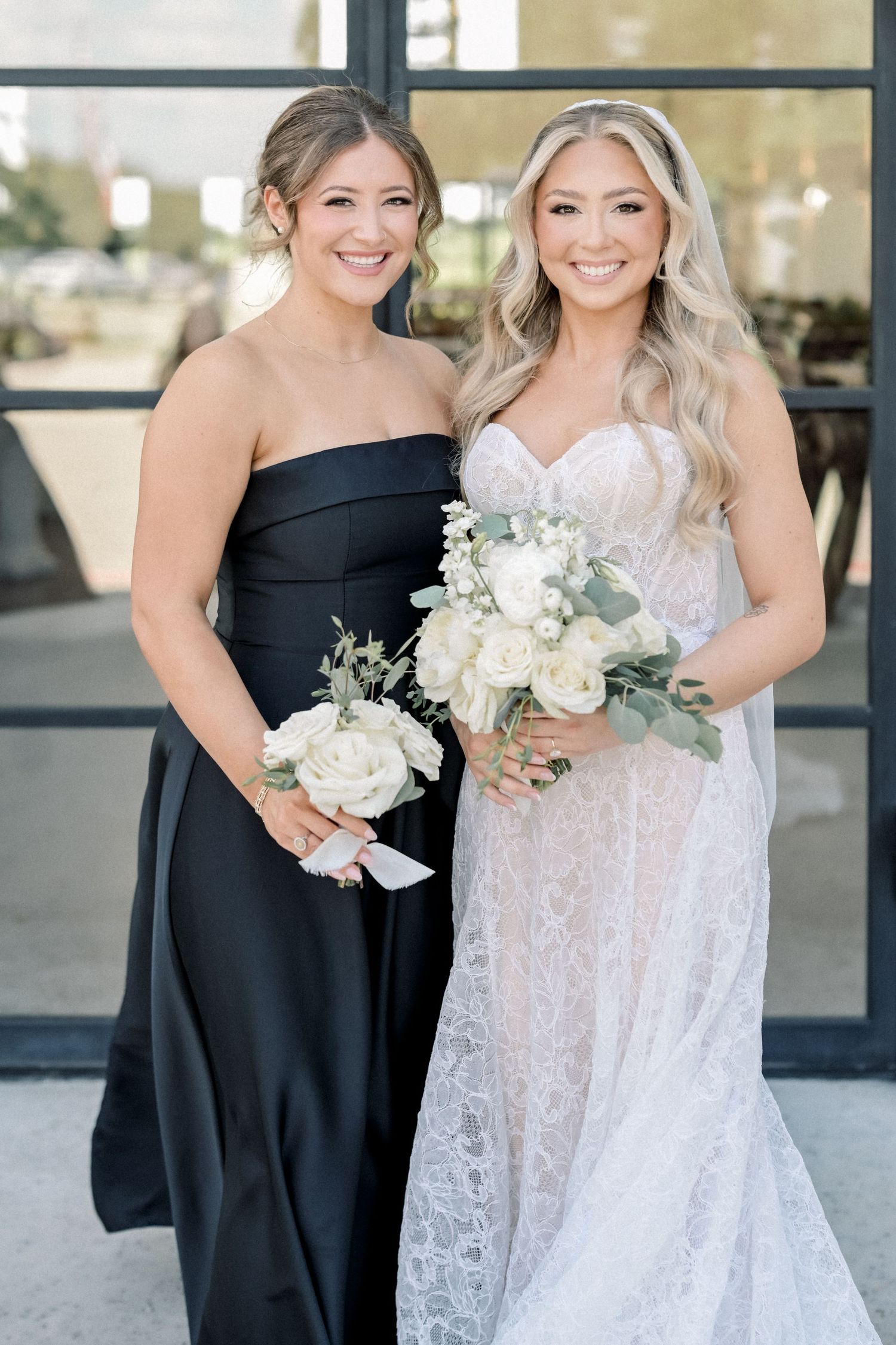 Bride in strapless lace gown and bridesmaid in black strapless dress smile holding white bouquets.