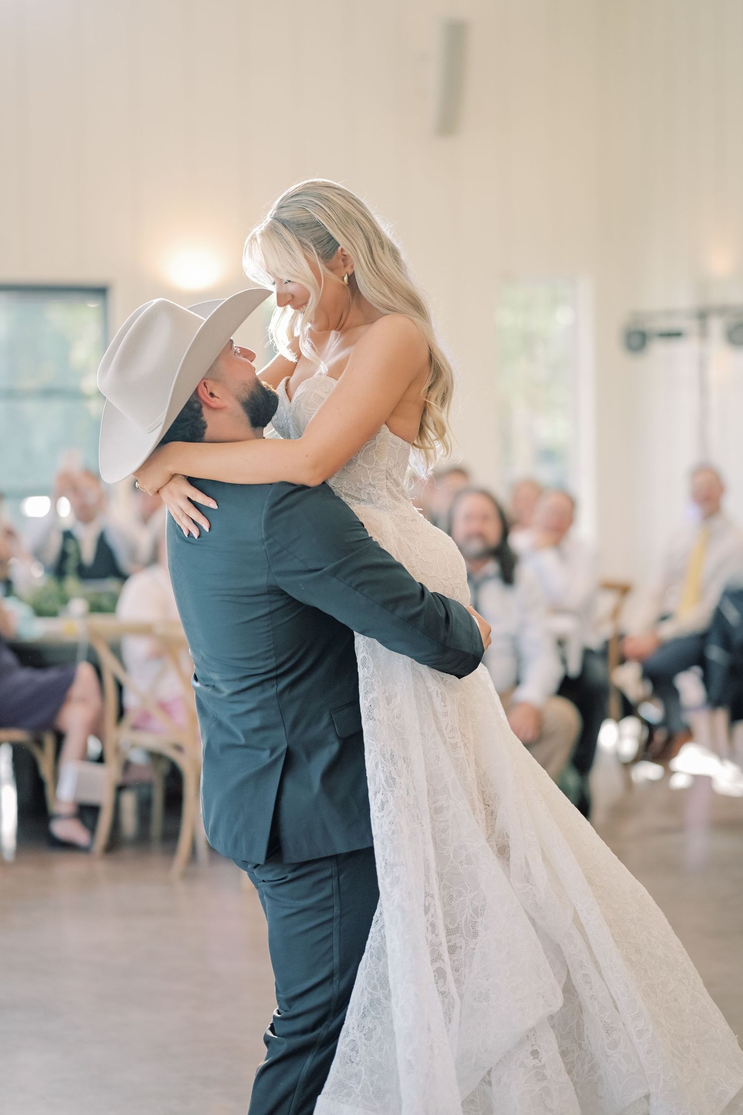 Groom in cowboy hat lifts smiling bride in lace gown during joyful first dance at wedding reception.