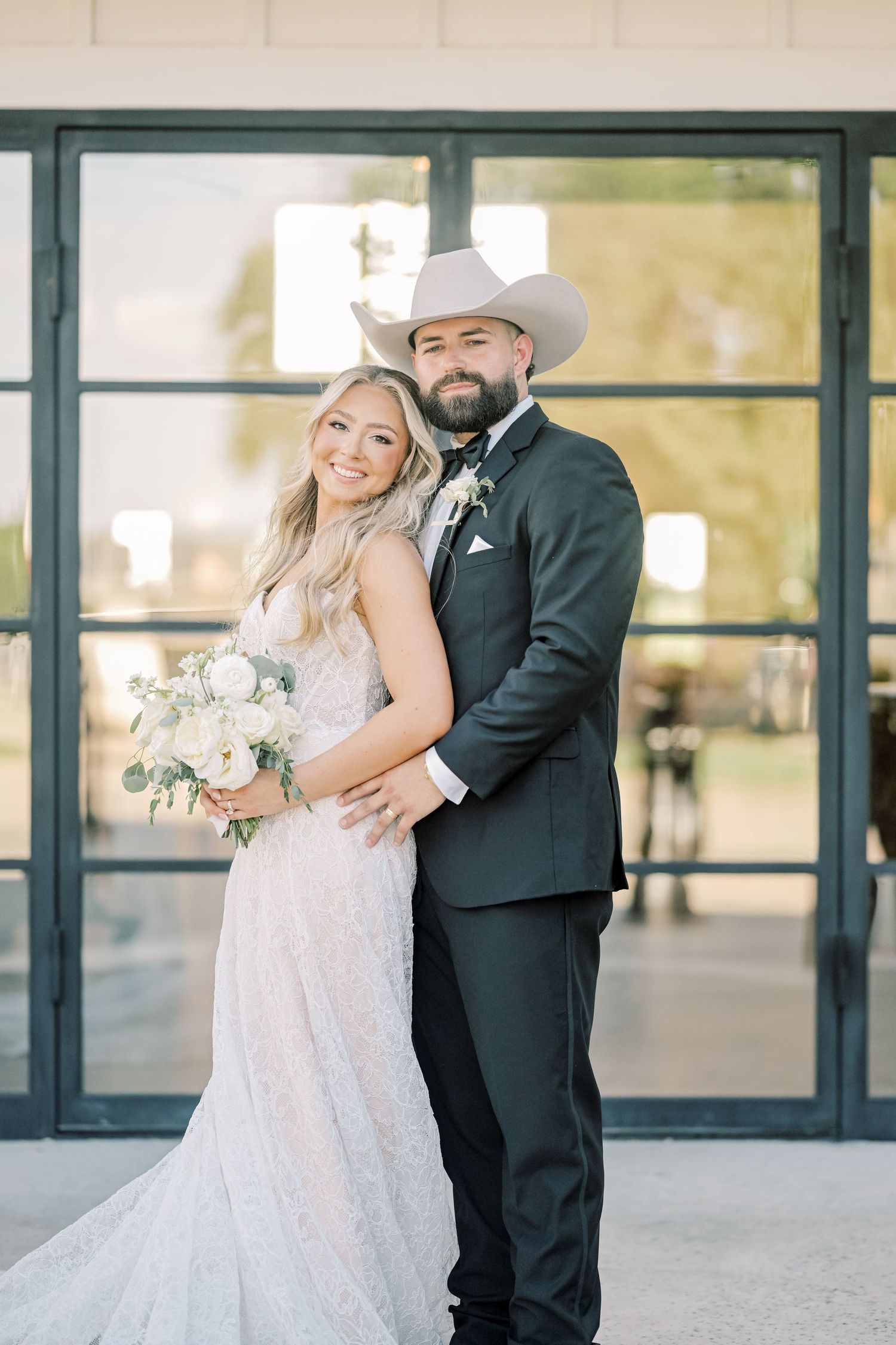Smiling bride and groom pose together outside venue, groom in cowboy hat holds bride close.
