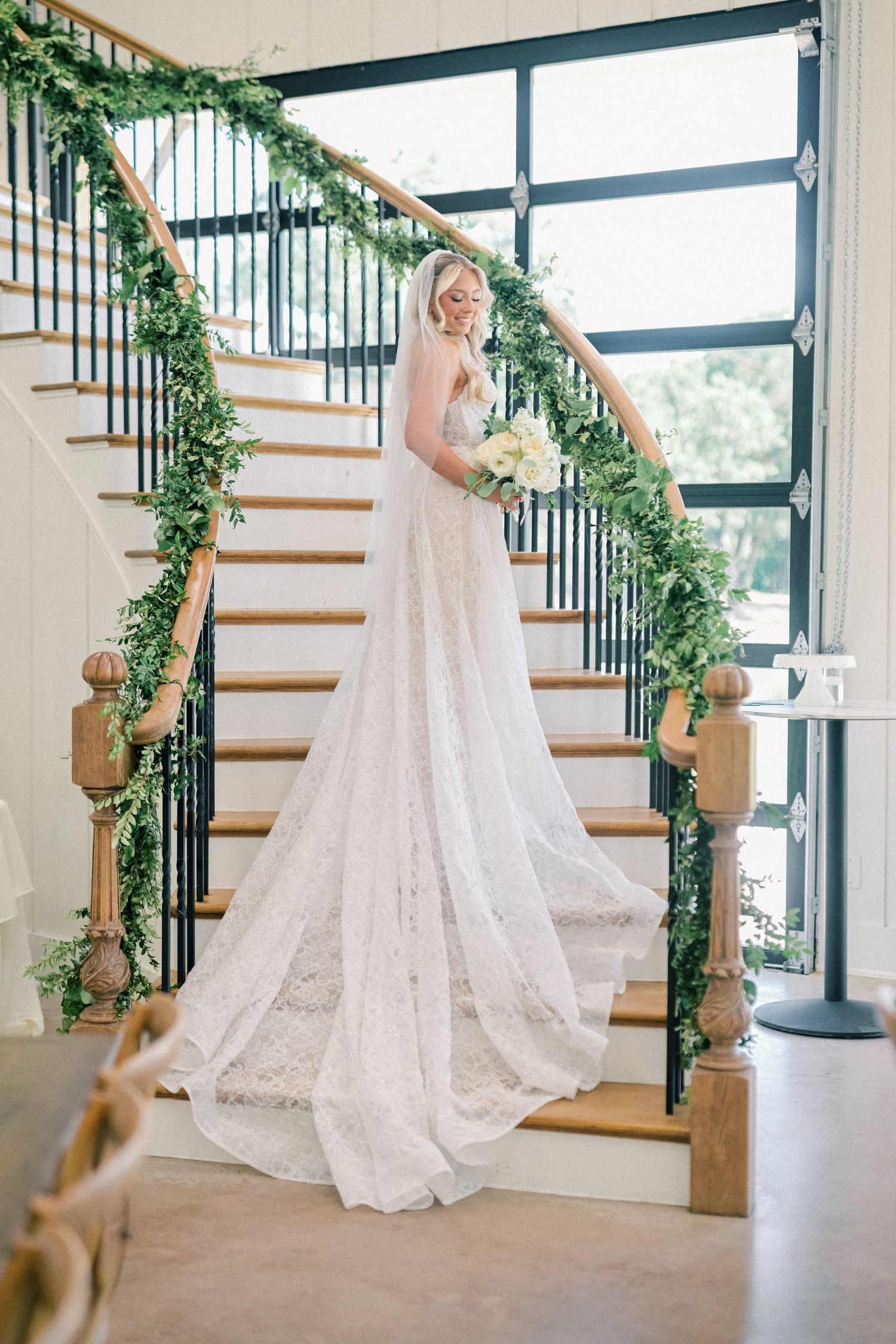 Elegant bride in lace wedding gown stands on staircase adorned with greenery garland holding white bouquet.