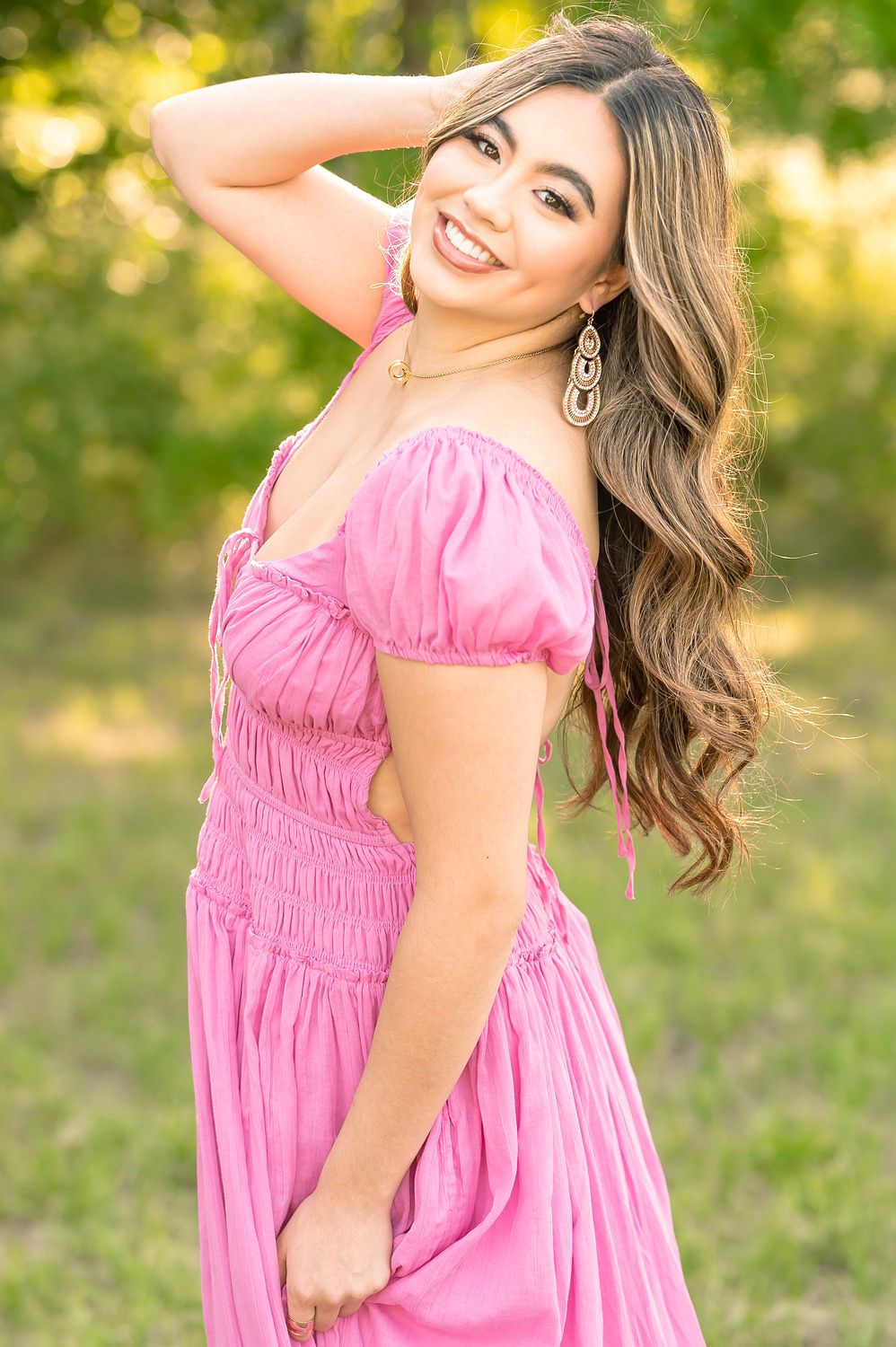 A young woman in a pink off-shoulder midi dress smiles outdoors, with long wavy hair and dangling earrings.