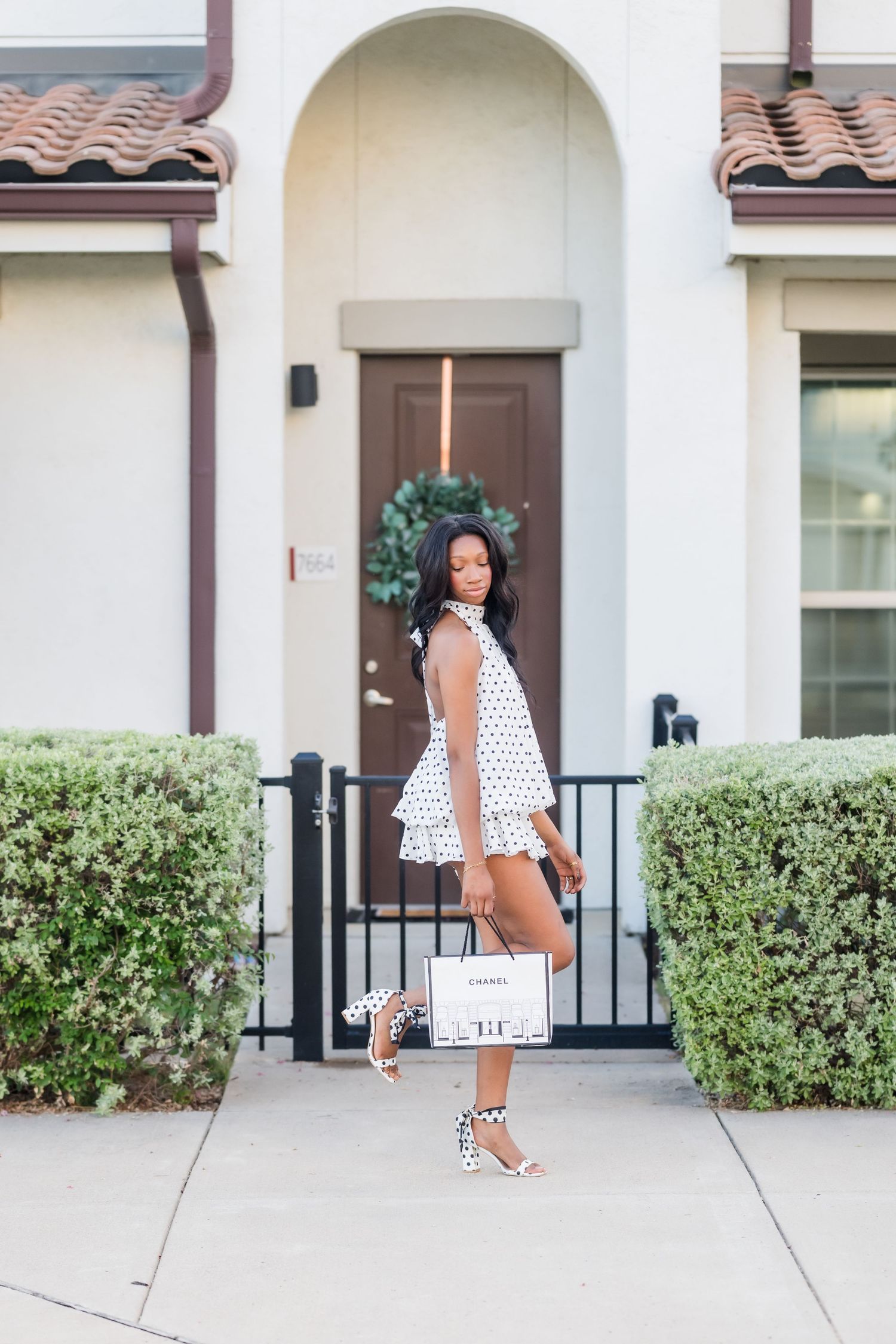 Woman in polka dot dress holding Chanel bag, posing on sidewalk in front of elegant home with wreath on door.