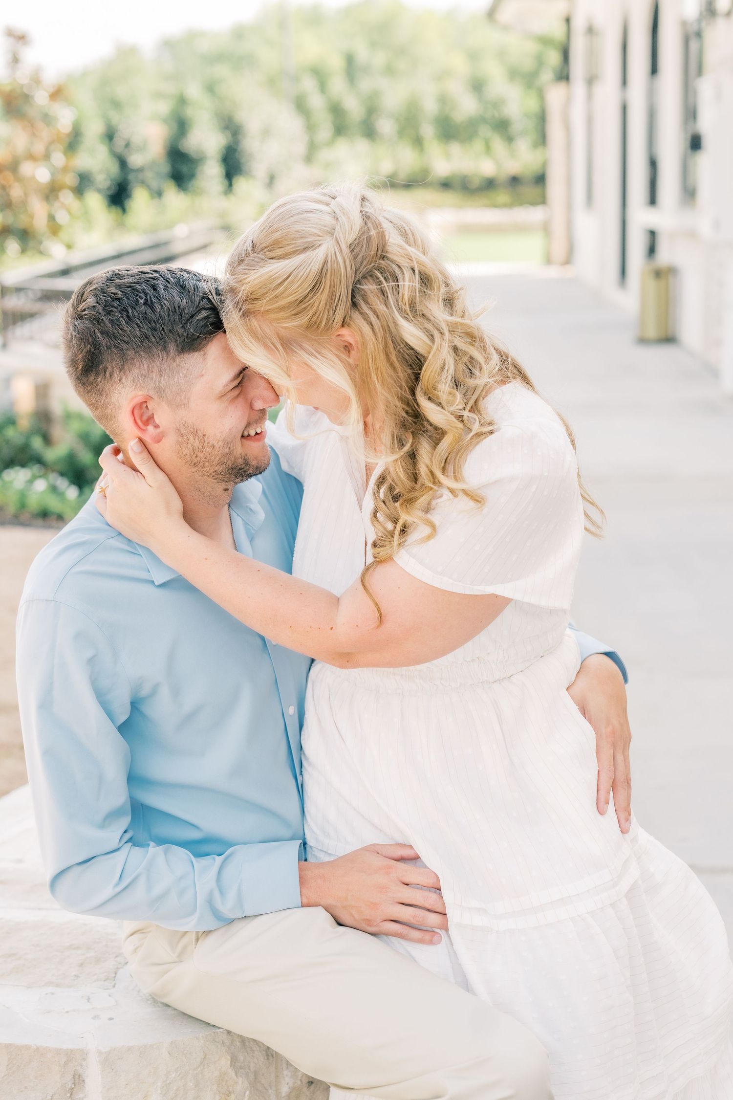 A couple shares a tender moment, the woman in a white dress cradling the mans face as they lean foreheads together outdoors.