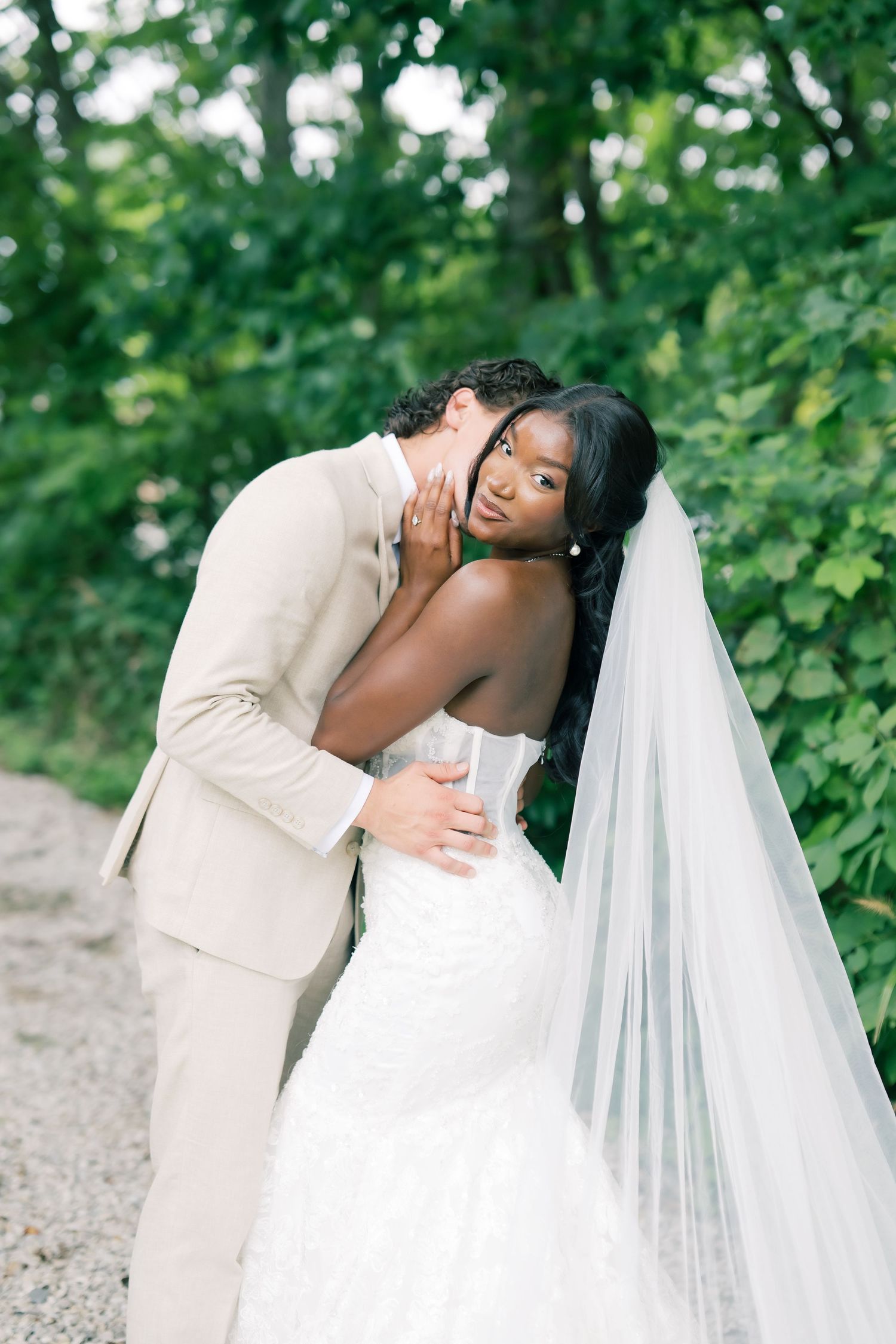A bride in a white mermaid gown and veil embraces her groom in a tan suit outdoors among lush greenery.