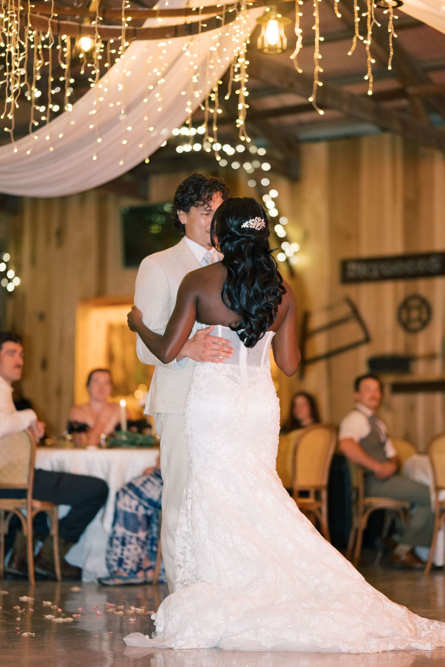 Bride and groom embrace during first dance under twinkling fairy lights at elegant barn reception.