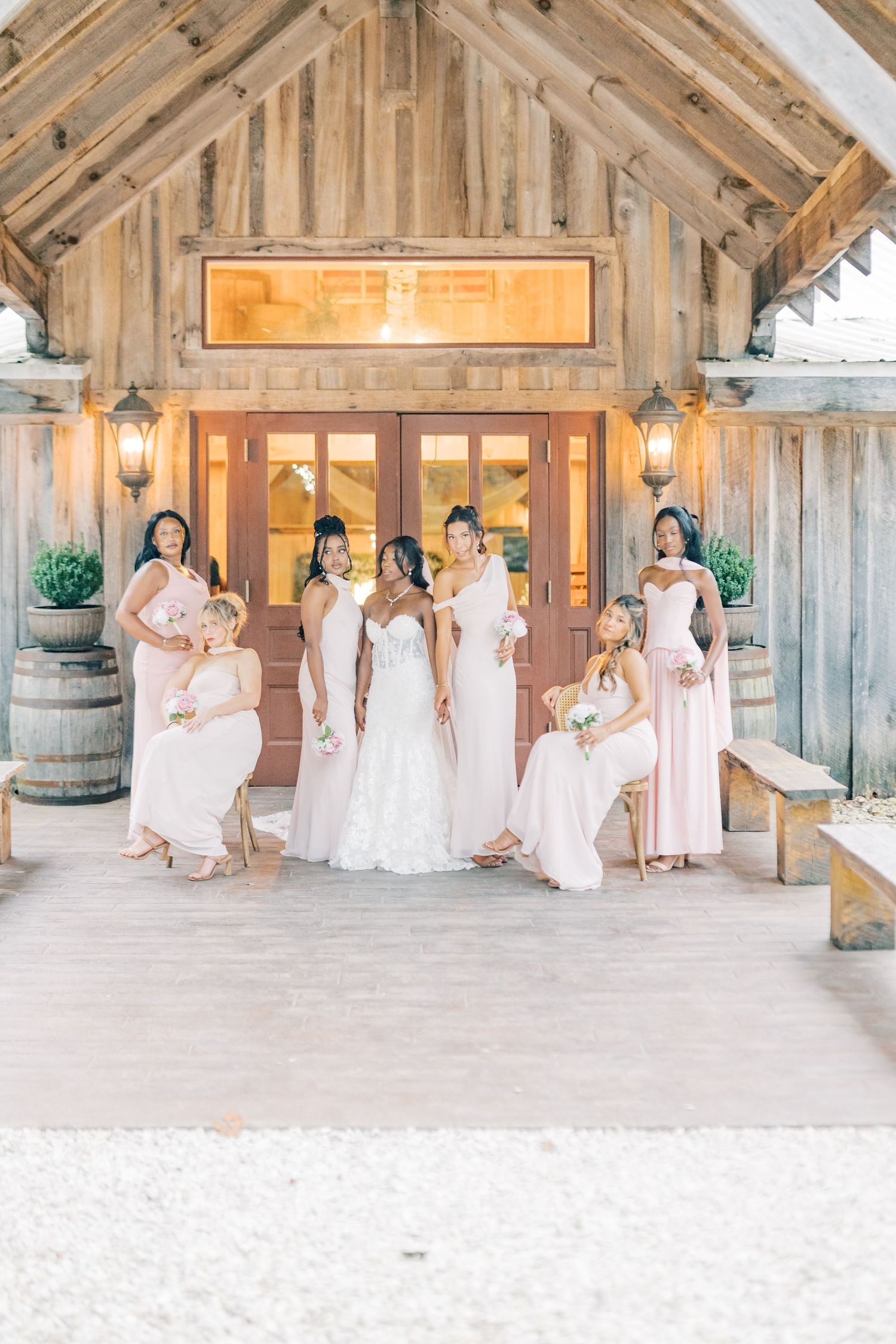 Bride in white gown stands with bridesmaids in blush dresses outside rustic barn wedding venue entrance.