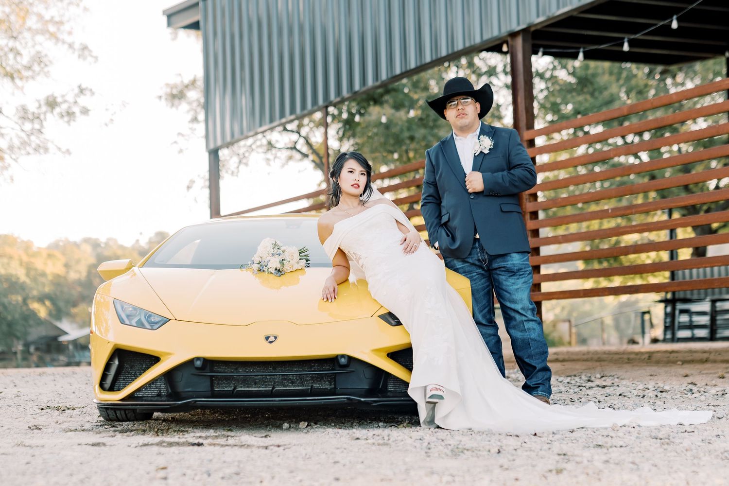 Bride and groom posing with a yellow Lamborghini at a rustic outdoor wedding venue at golden hour.