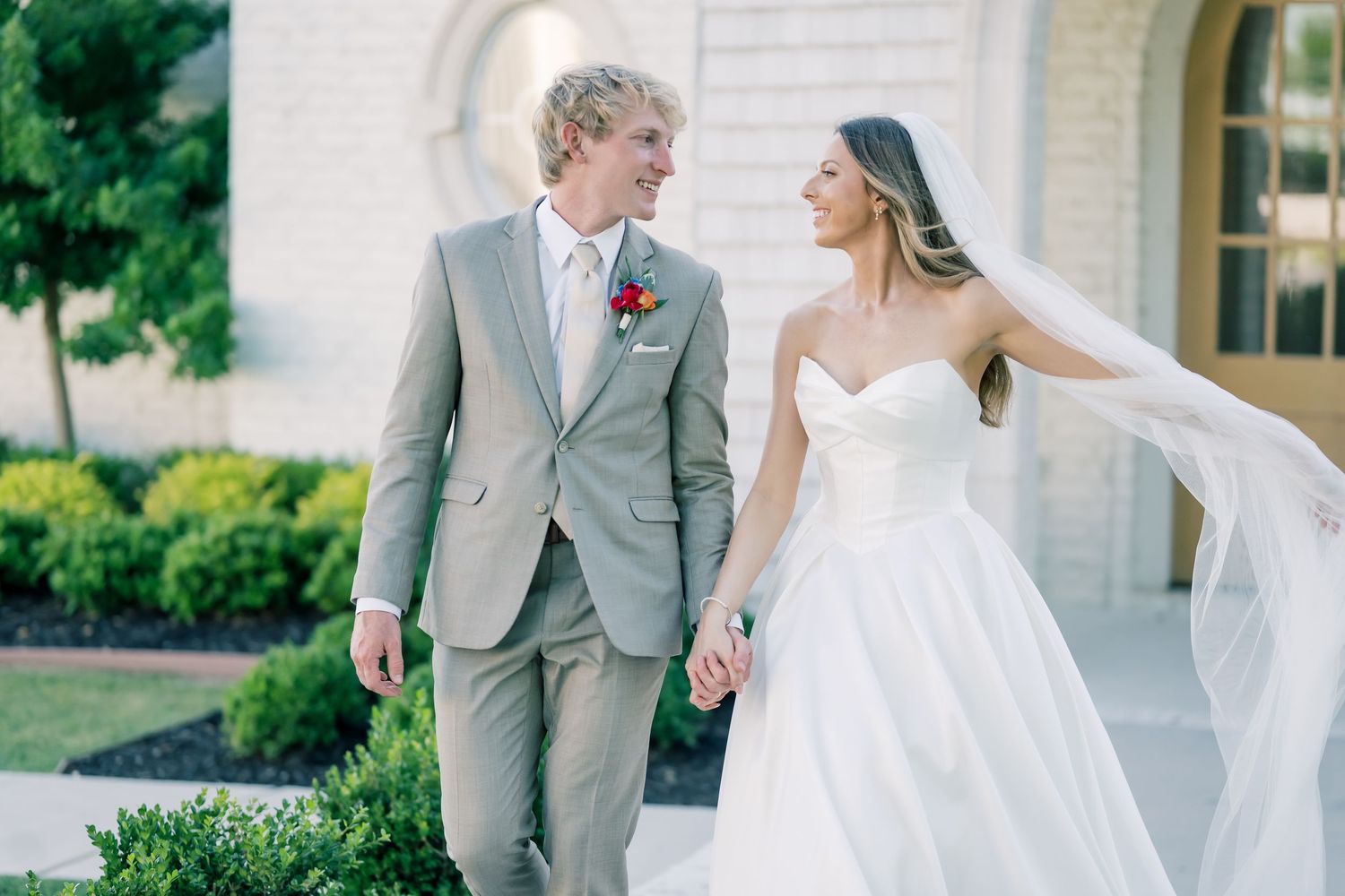 Bride in white strapless gown with flowing veil holds hands with groom in tan suit with boutonniere outdoors.