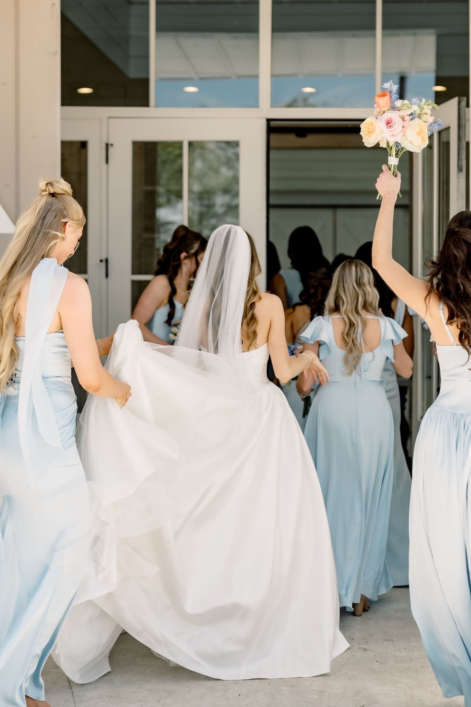 Bride in white gown and veil walks with bridesmaids in light blue dresses toward venue entrance, bouquet raised.