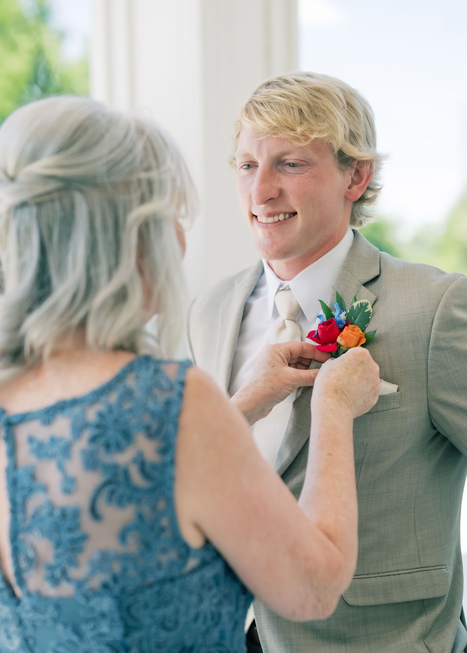 Mother pinning colorful boutonniere on smiling groom in gray suit at wedding preparation.