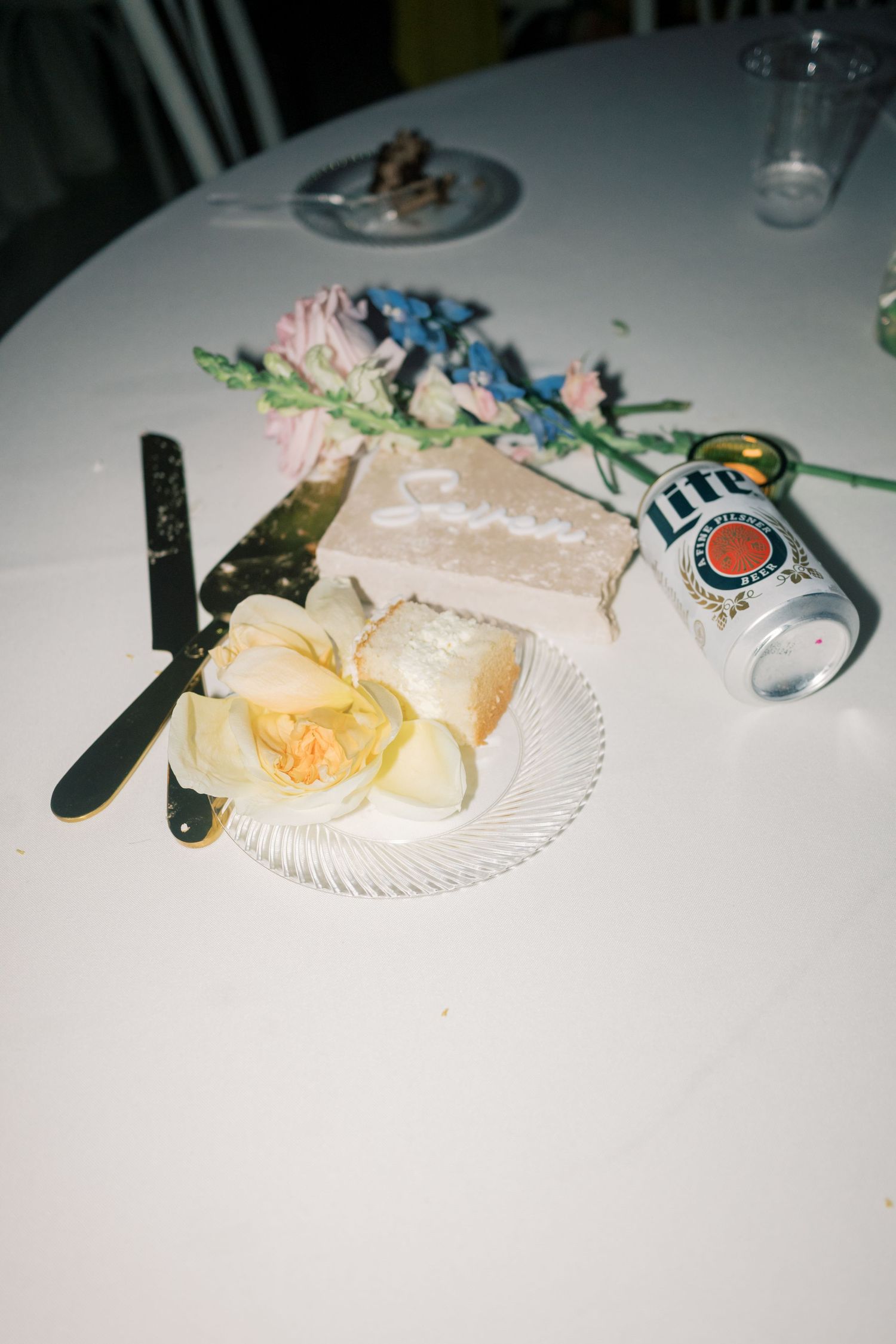 Wedding cake slice on plate with orchid, Miller Lite can, knife, and floral arrangement on white table.