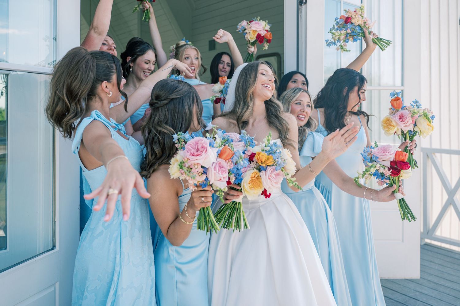 Bride in white gown with bridesmaids in light blue dresses celebrating with colorful bouquets at outdoor wedding.