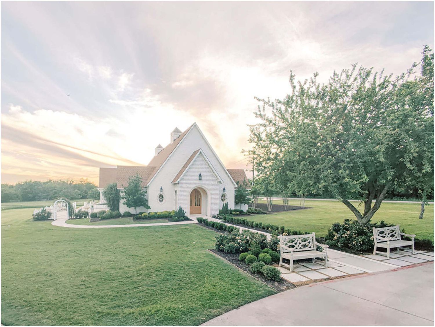 White chapel wedding venue with manicured gardens, white benches, and a large tree at sunset.