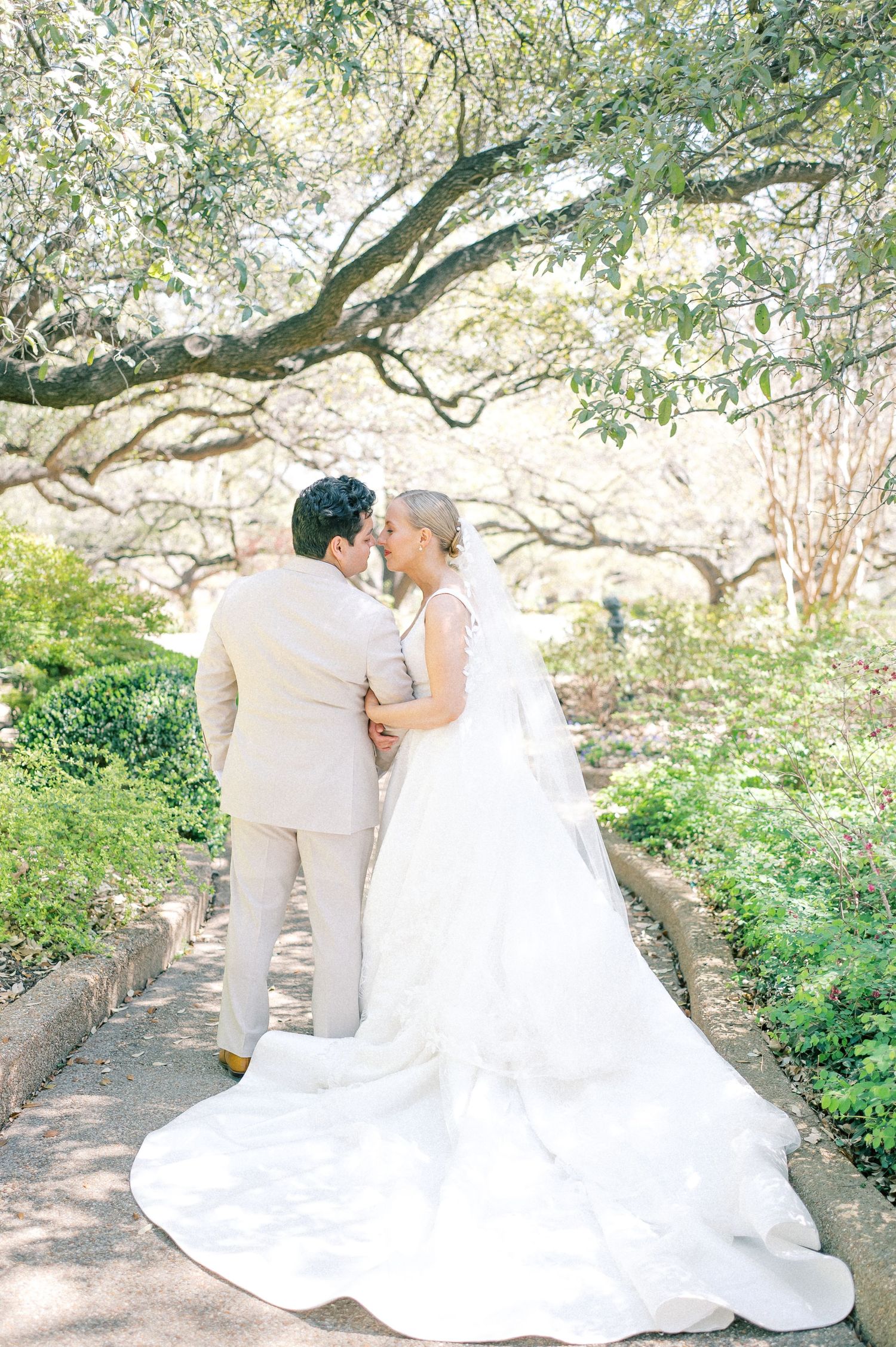 A romantic wedding couple embraces under a sprawling tree in a garden setting.