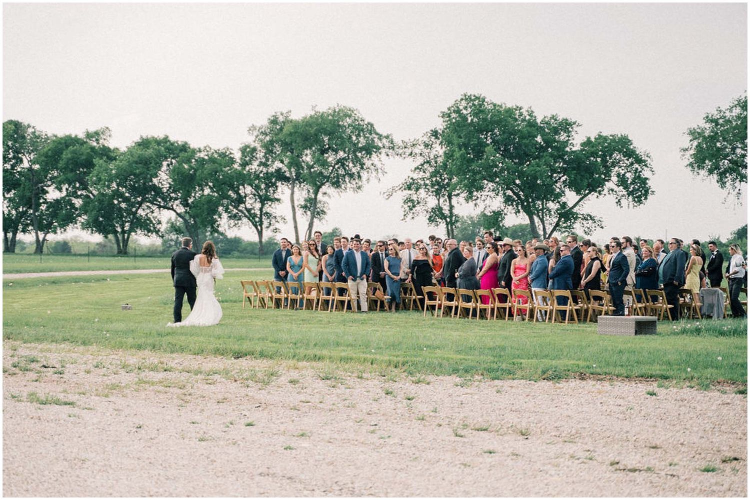 Outdoor wedding ceremony with guests seated on wooden chairs in a grassy field.