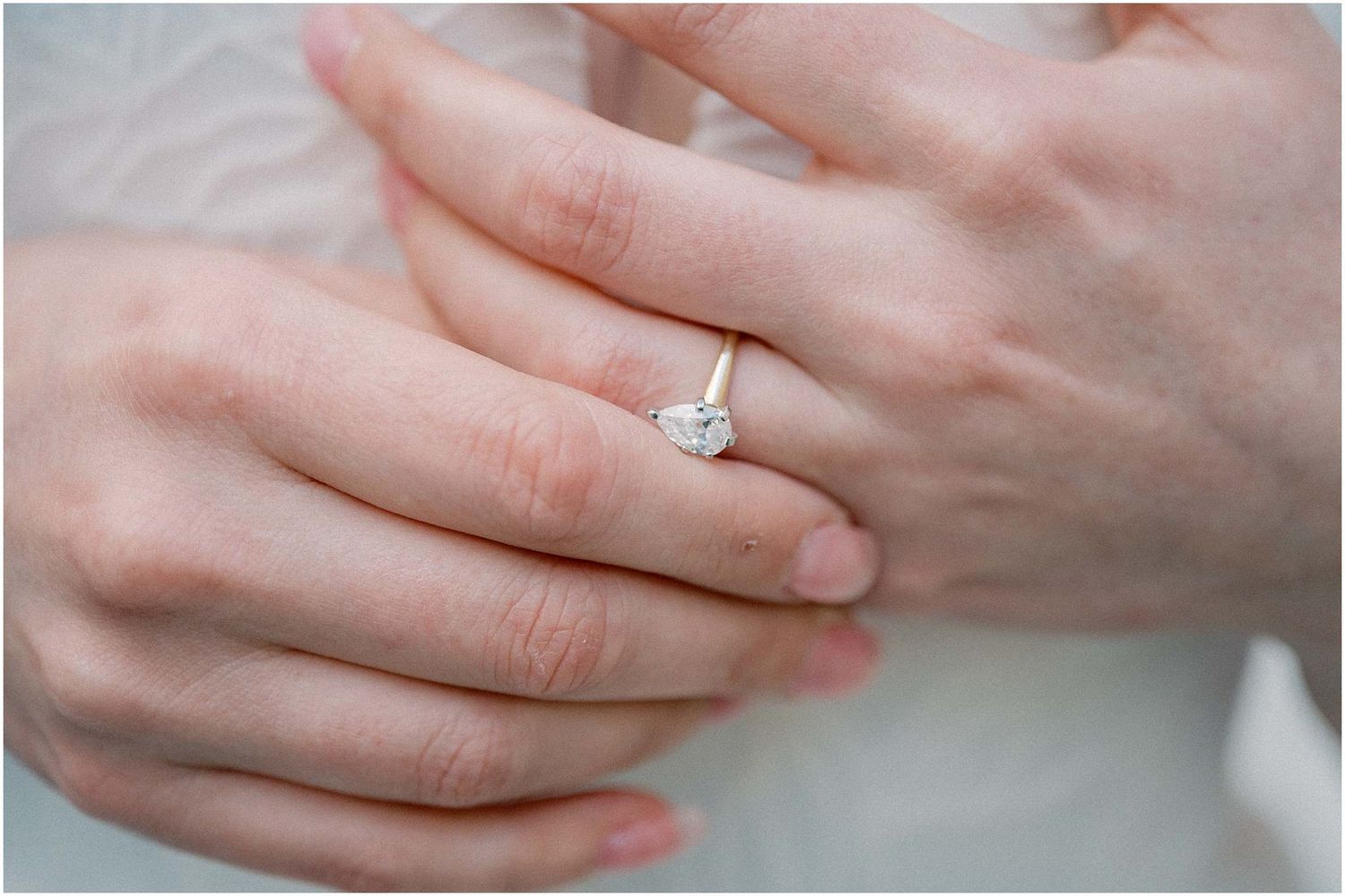 Close-up of engagement ring and delicate hands against white fabric.