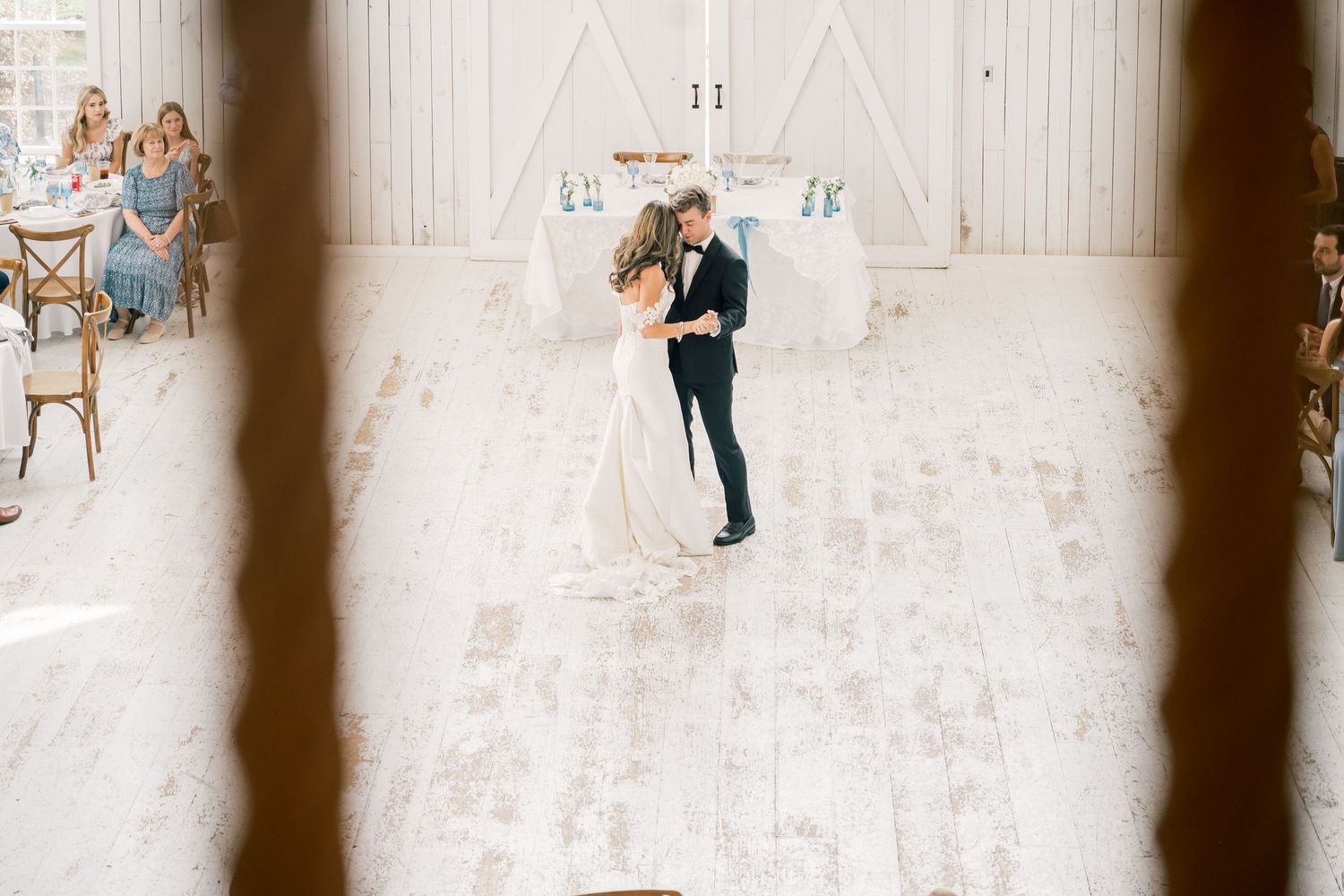 Bride and groom share first dance in white barn wedding venue with guests seated.