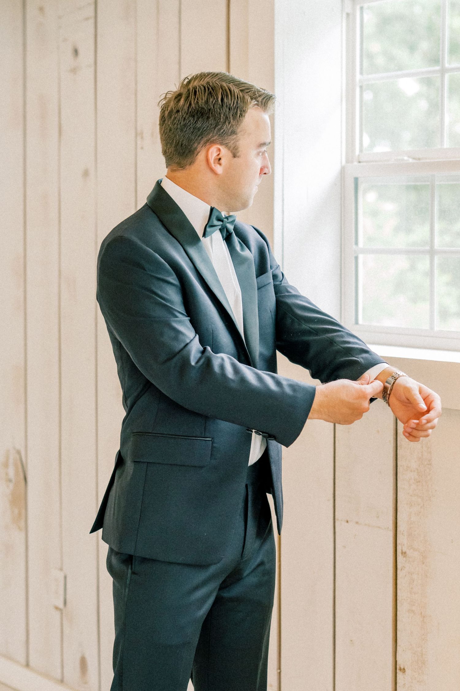Dapper groom in navy suit and teal bow tie adjusting watch by window.