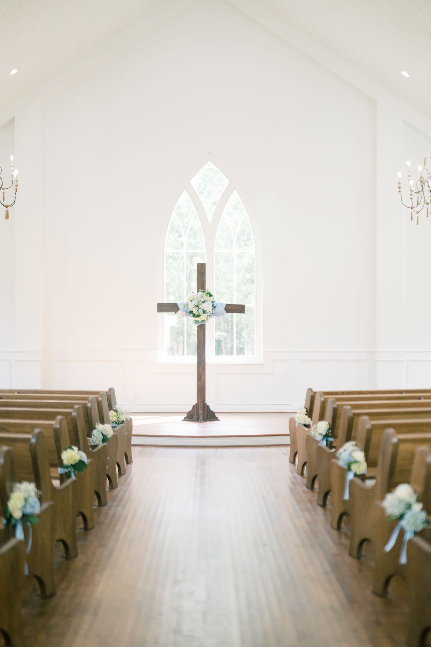 Elegant white chapel interior with wooden cross, pews adorned with floral arrangements and chandelier lighting.
