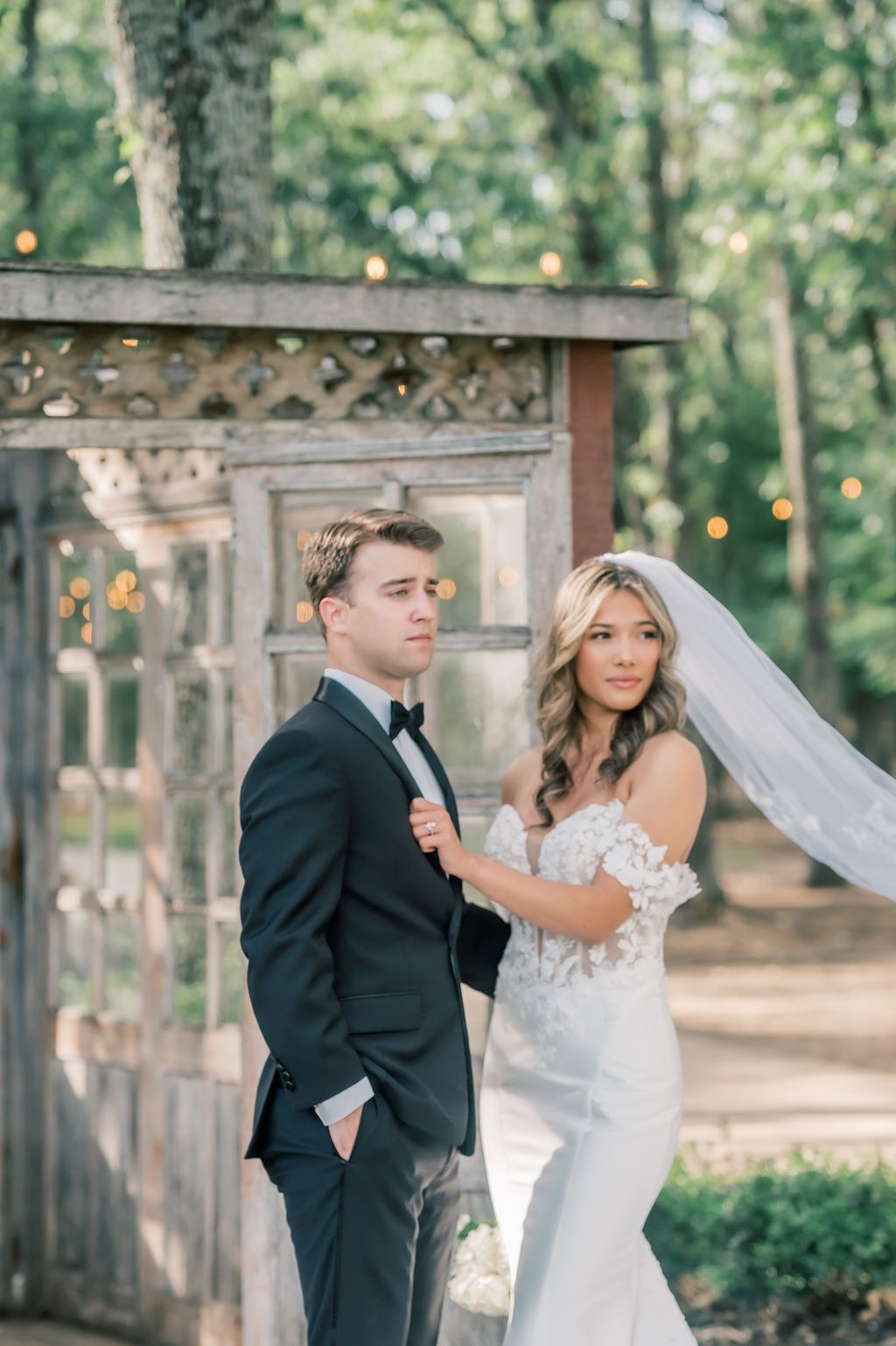 Bride in lace gown and veil stands with groom in black tuxedo before rustic wooden arbor in wooded outdoor setting.