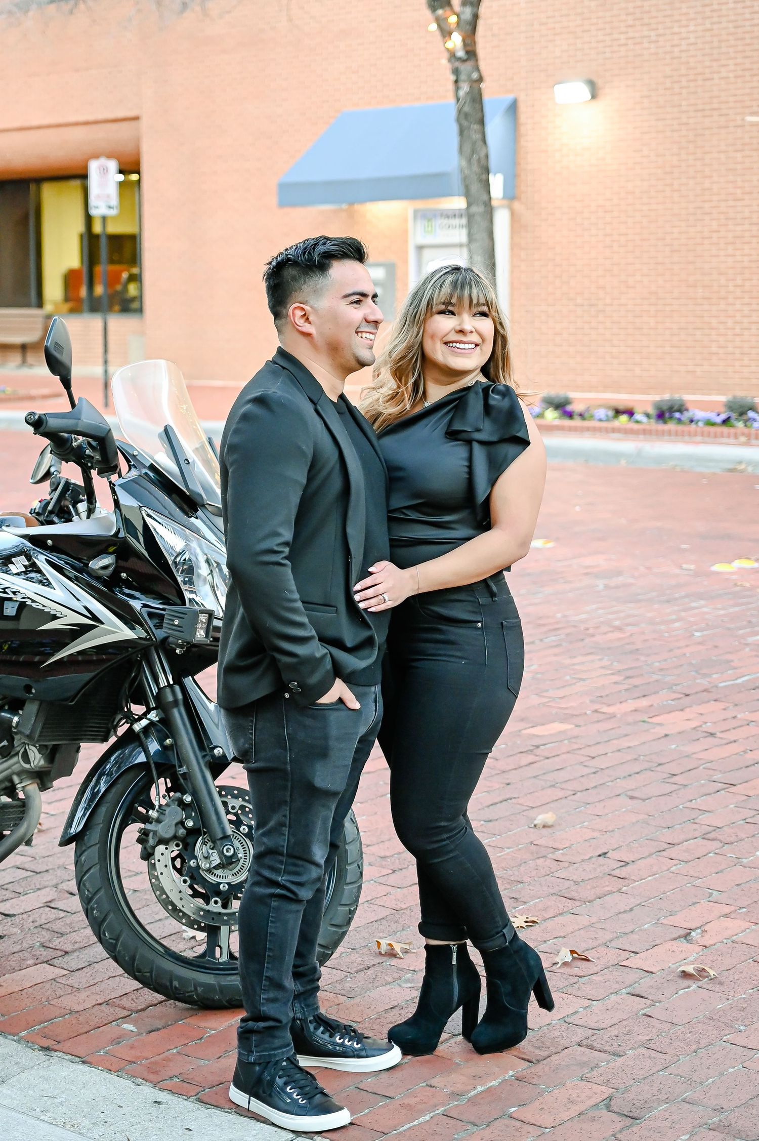 Couple dressed in all-black outfits posing together on a brick sidewalk near a motorcycle in an urban setting.