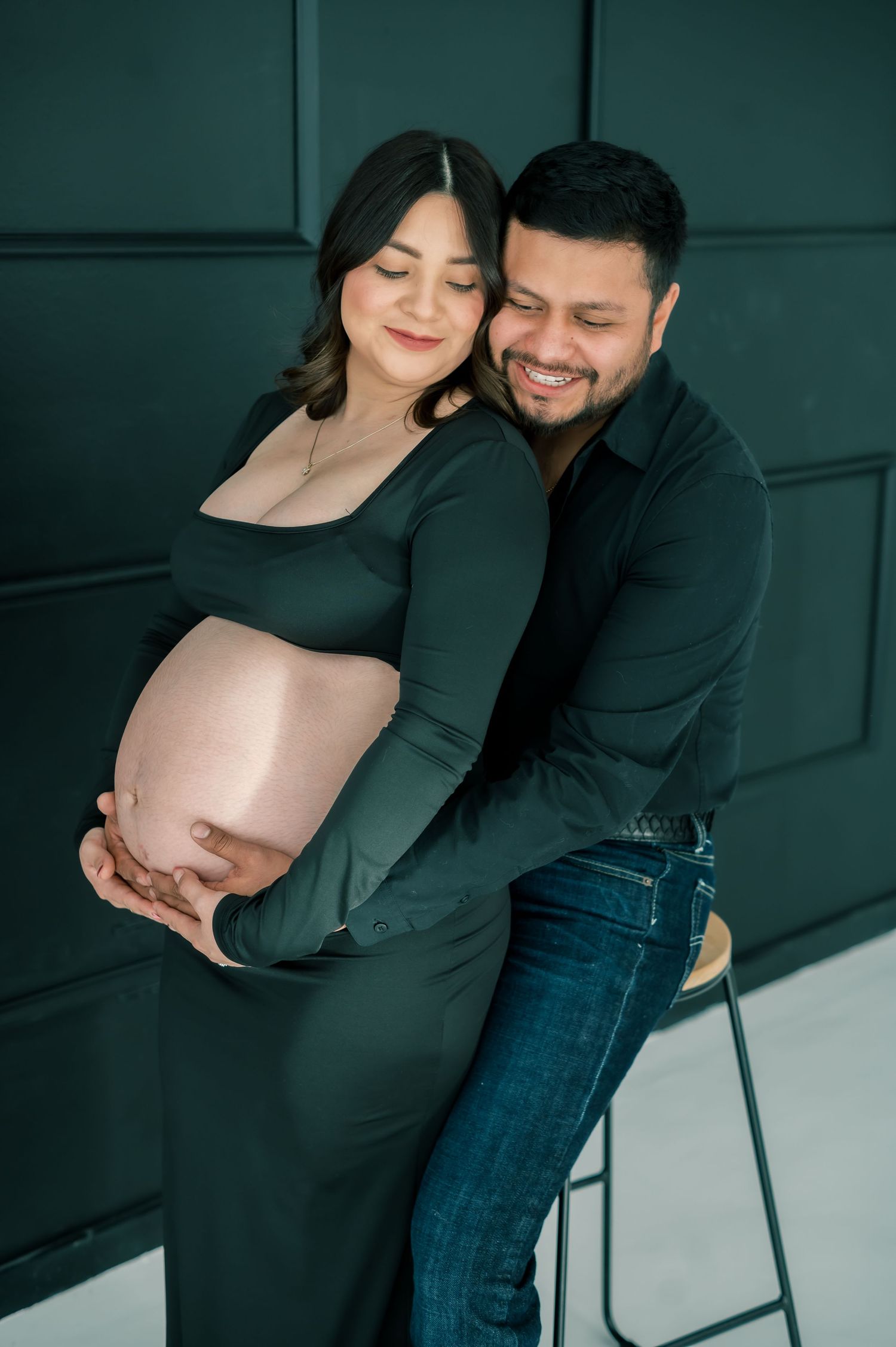 Smiling couple in black outfits embracing during maternity photo shoot against dark wall.